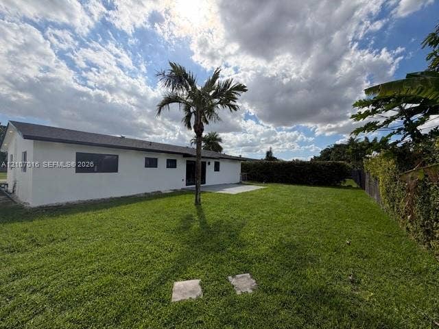 Rear exterior featuring white stucco siding, dark-toned roof shingles, rear concrete patio, expansive grass lawn, and mature palm tree