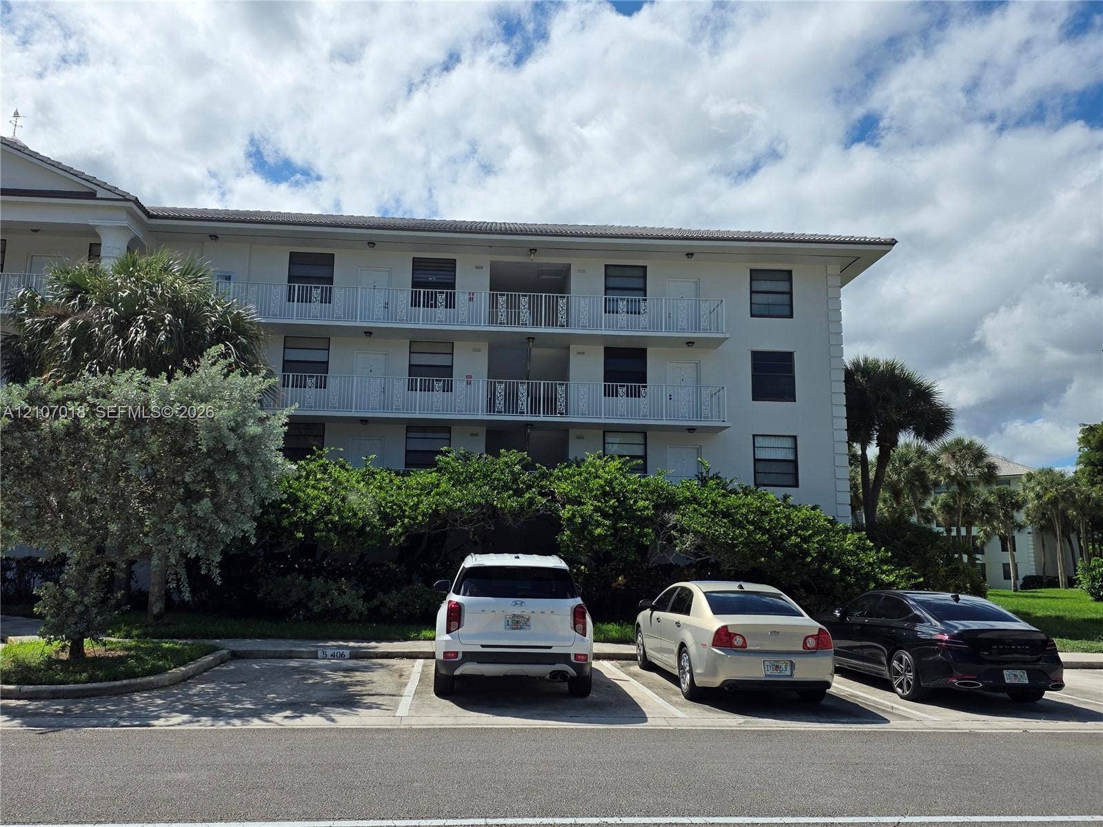 3716 Whitehall Drive, #304, West Palm Beach, West Palm Beach, FL 33401 Multi-story residential building exterior featuring a white stucco facade, shingle roof, integrated metal balcony railings, and assigned surface parking spaces