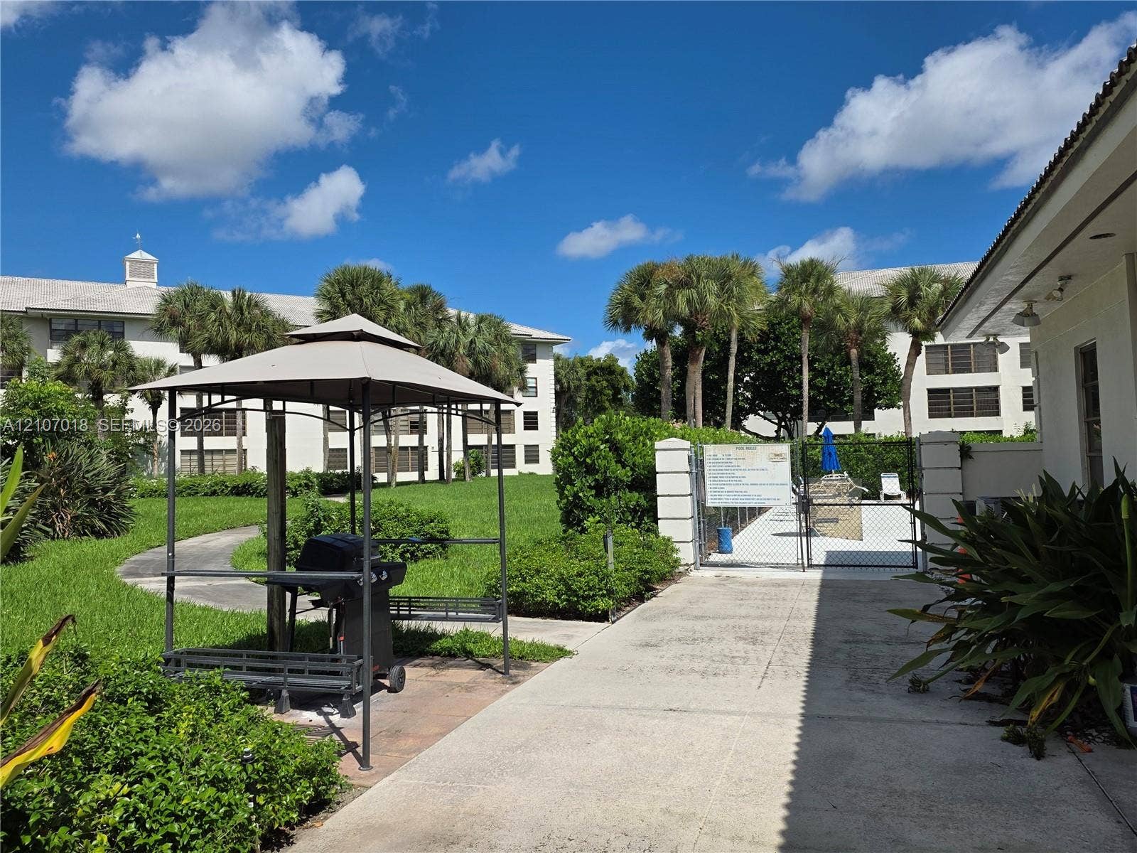 3716 Whitehall Drive, #304, West Palm Beach, West Palm Beach, FL 33401 Outdoor common area featuring a paved walkway, manicured green space, mature palm trees, and a covered grilling station