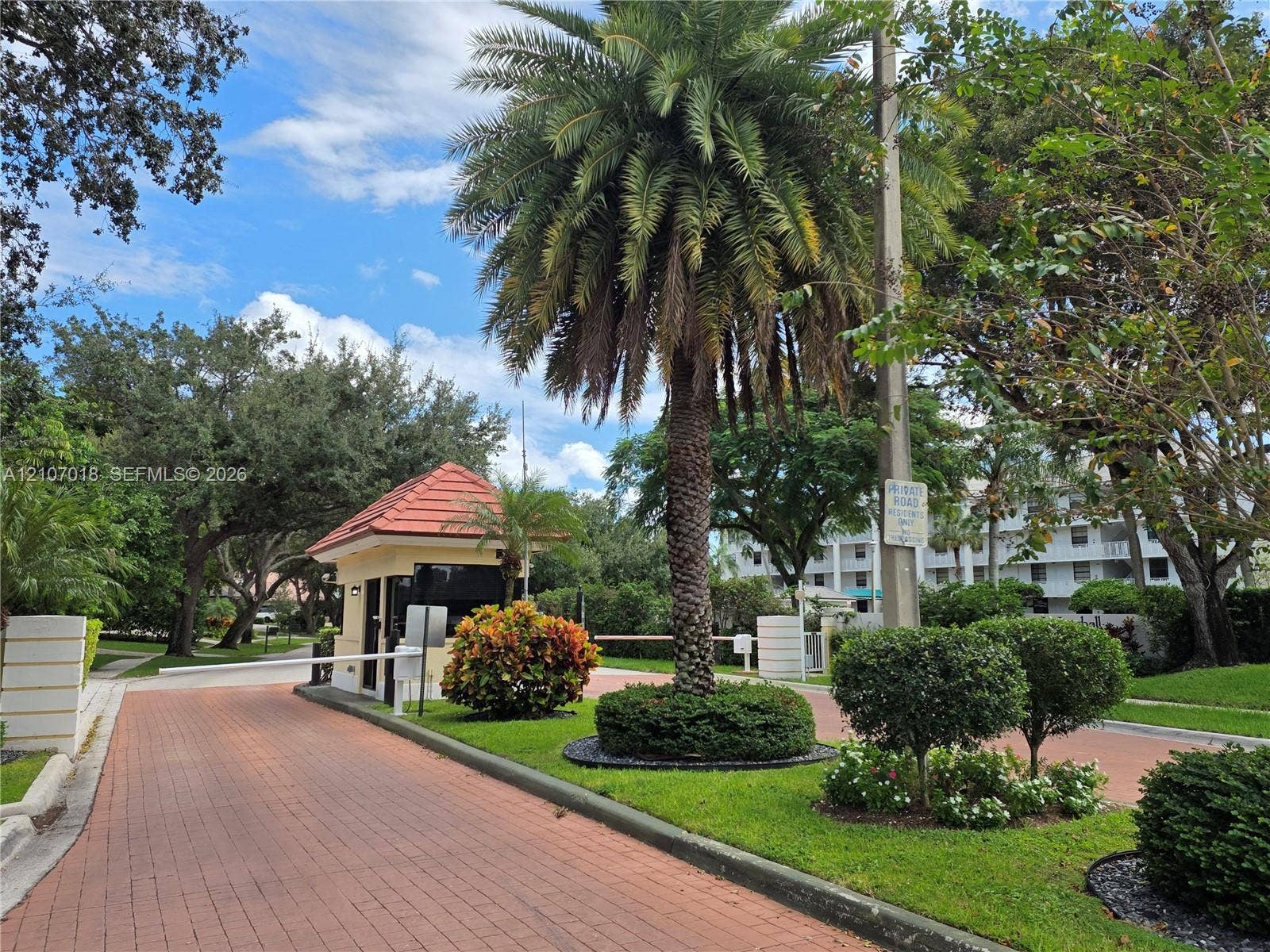 3716 Whitehall Drive, #304, West Palm Beach, West Palm Beach, FL 33401 Gated entrance featuring a brick-paver roadway, guard booth with a tile roof, mature palm trees, and manicured landscaping