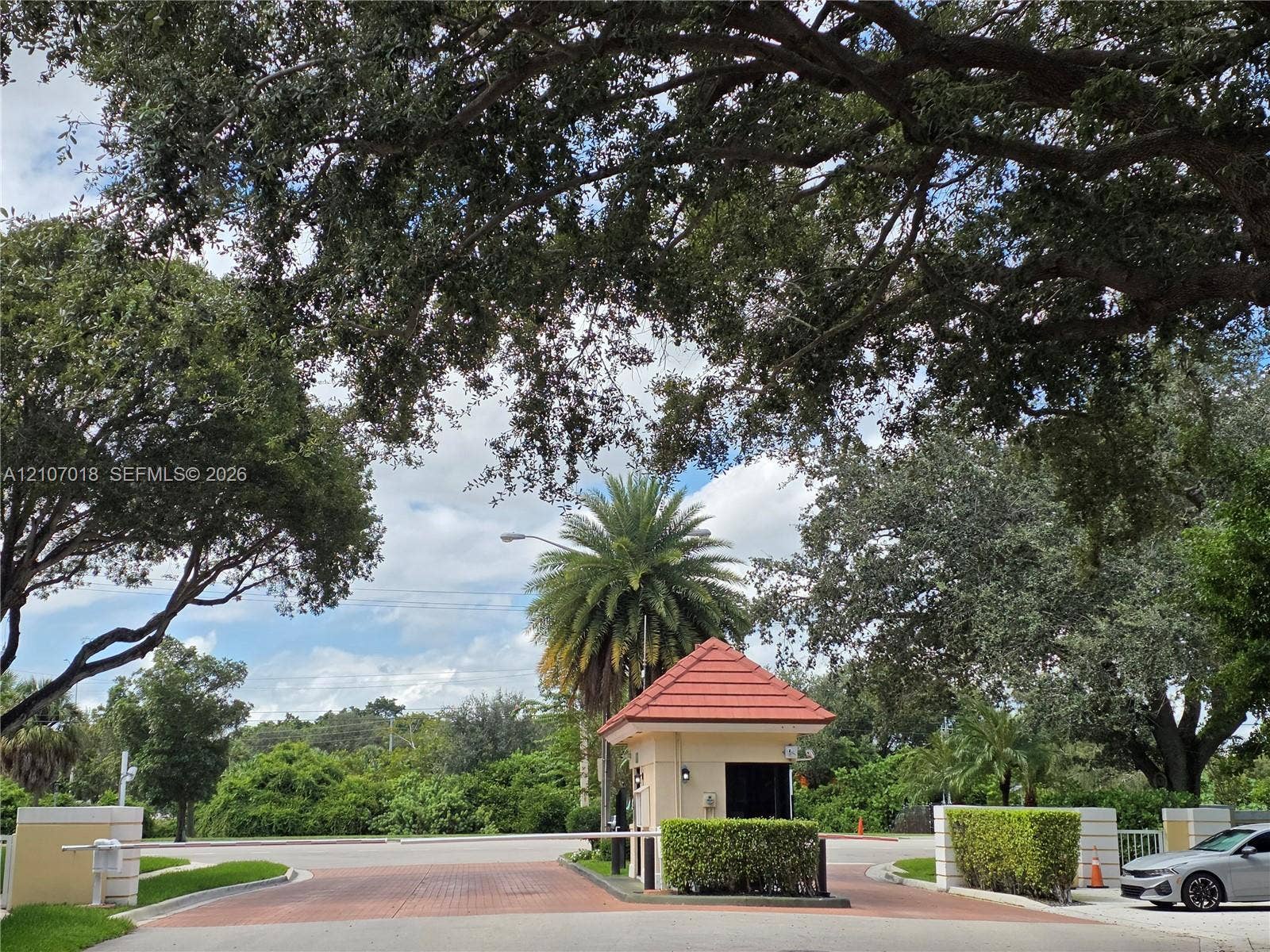 3716 Whitehall Drive, #304, West Palm Beach, West Palm Beach, FL 33401 Gated entrance featuring a guardhouse with a tiled roof, paved brick thoroughfare, and mature landscape canopy