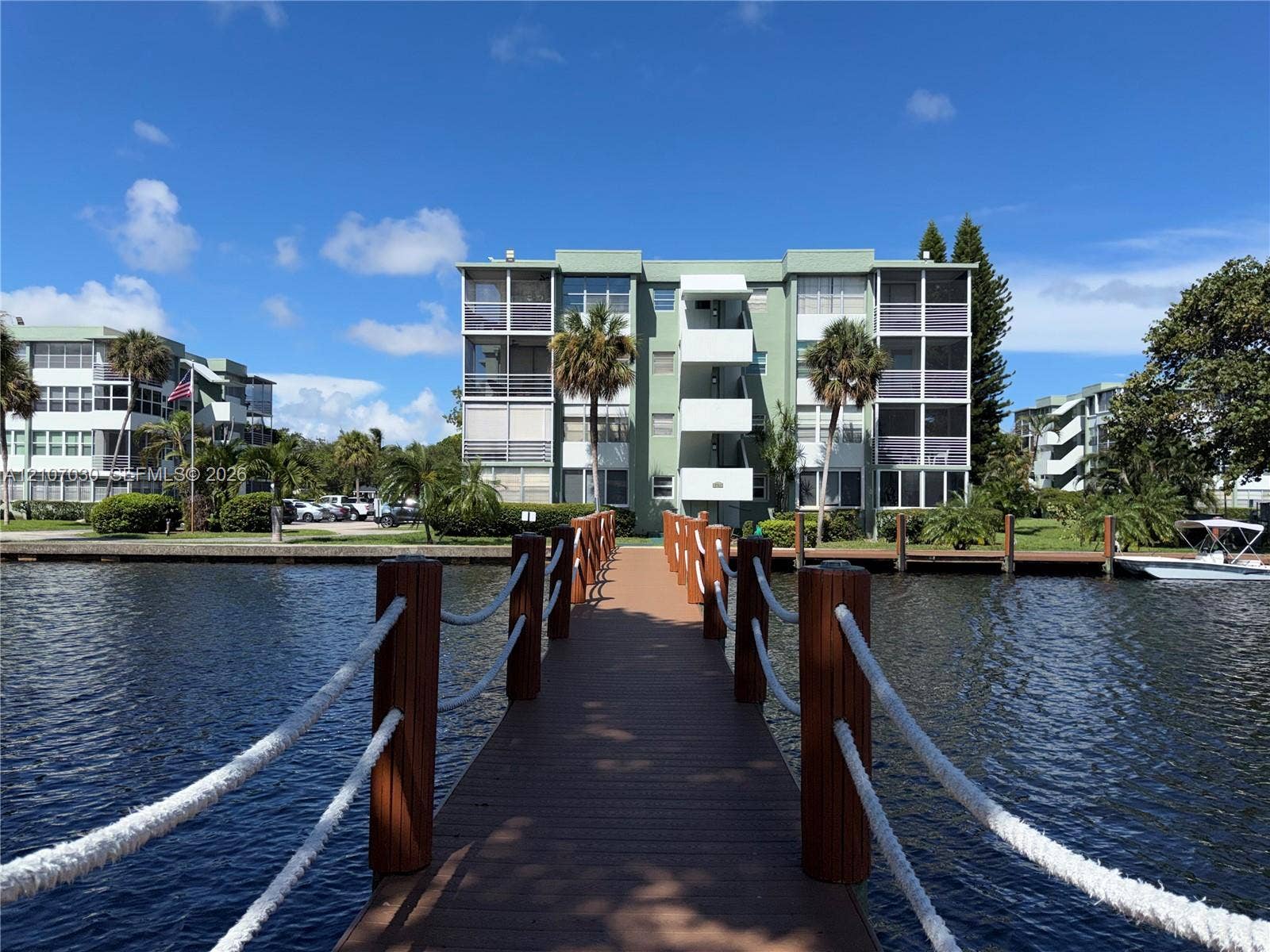 Waterfront dock featuring synthetic decking, wood posts, and nautical rope railings leading to a multi-story building with screened balconies and exterior walkways