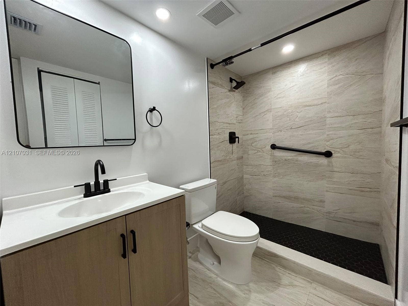 Bathroom featuring a wood-finish vanity with a white countertop, matte black fixtures, a rectangular vanity mirror with rounded corners, and a walk-in shower with stone-patterned wall tile and a black hexagonal tile floor