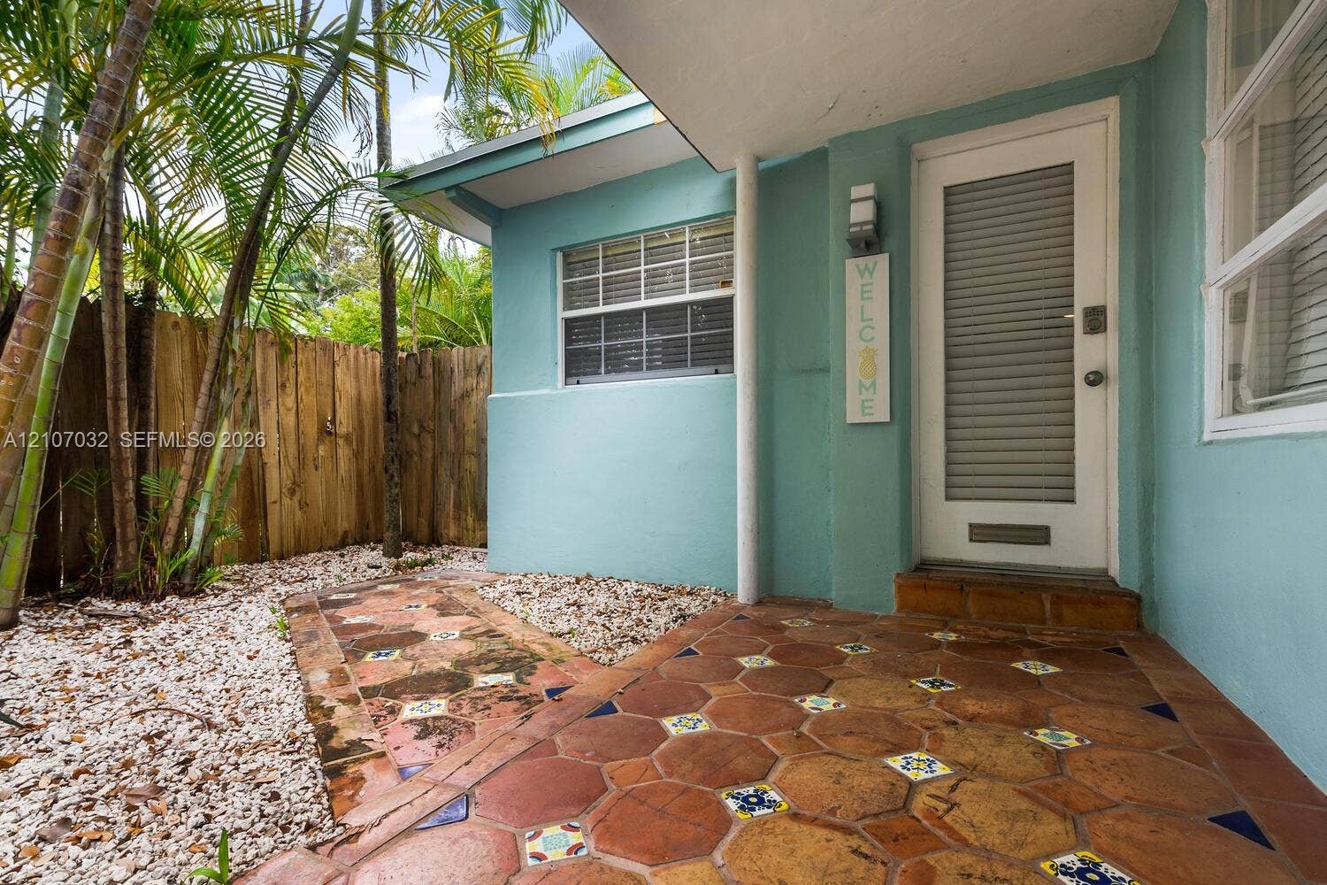 443 NE 17th Way, Fort Lauderdale, FL 33301 Entryway featuring a patterned terracotta tile porch, louvered glass door, smooth-finish exterior siding, stone-ground landscaping, and a natural wood privacy fence