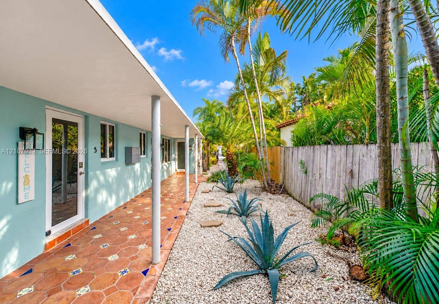443 NE 17th Way, Fort Lauderdale, FL 33301 Covered walkway featuring terra cotta tile flooring with decorative inlay, light-blue exterior walls, support columns, and xeriscaped garden beds with stone accents and mature palm trees