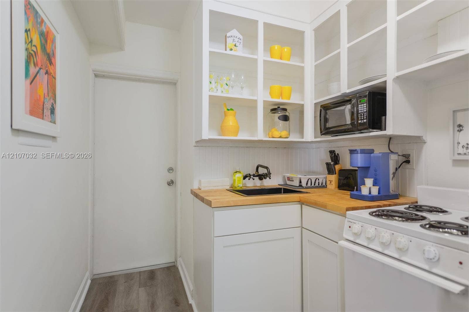 443 NE 17th Way, Fort Lauderdale, FL 33301 Kitchenette featuring white shaker-style cabinetry, open shelving with glass-front uppers, butcher block countertops, a black undermount sink with a matte black faucet, and wood-finish flooring