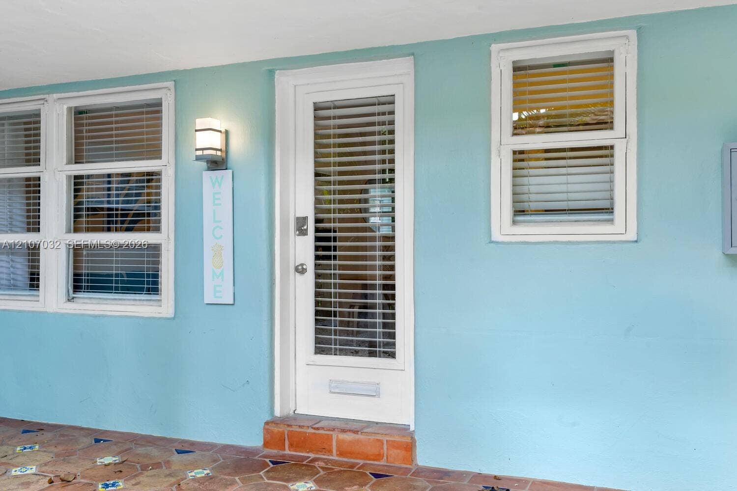 443 NE 17th Way, Fort Lauderdale, FL 33301 Entryway featuring a glass-panel door with a built-in mail slot, white-framed horizontal windows with integrated blinds, a wall-mounted exterior light fixture, and terracotta-tile flooring