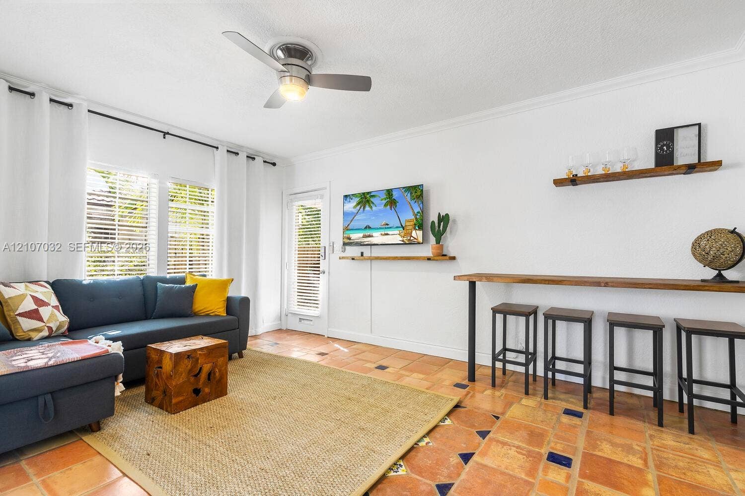 Living space featuring terracotta tile flooring with decorative tile inlays, a mounted ceiling fan, wall-mounted natural wood shelving, and a full-height glass-paneled exterior door