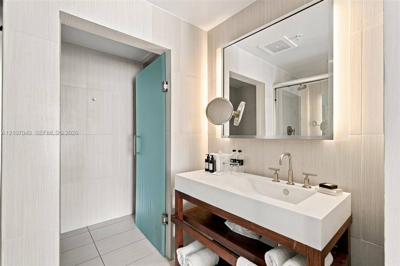 3101 Bayshore Drive, #707, Fort Lauderdale, FL 33304 Open-concept vanity featuring a white composite countertop, integrated rectangular sink, metal widespread faucet, wood-finish shelving, and wall-mounted mirror with integrated lighting, finished with floor-to-ceiling tile work and a frosted glass door