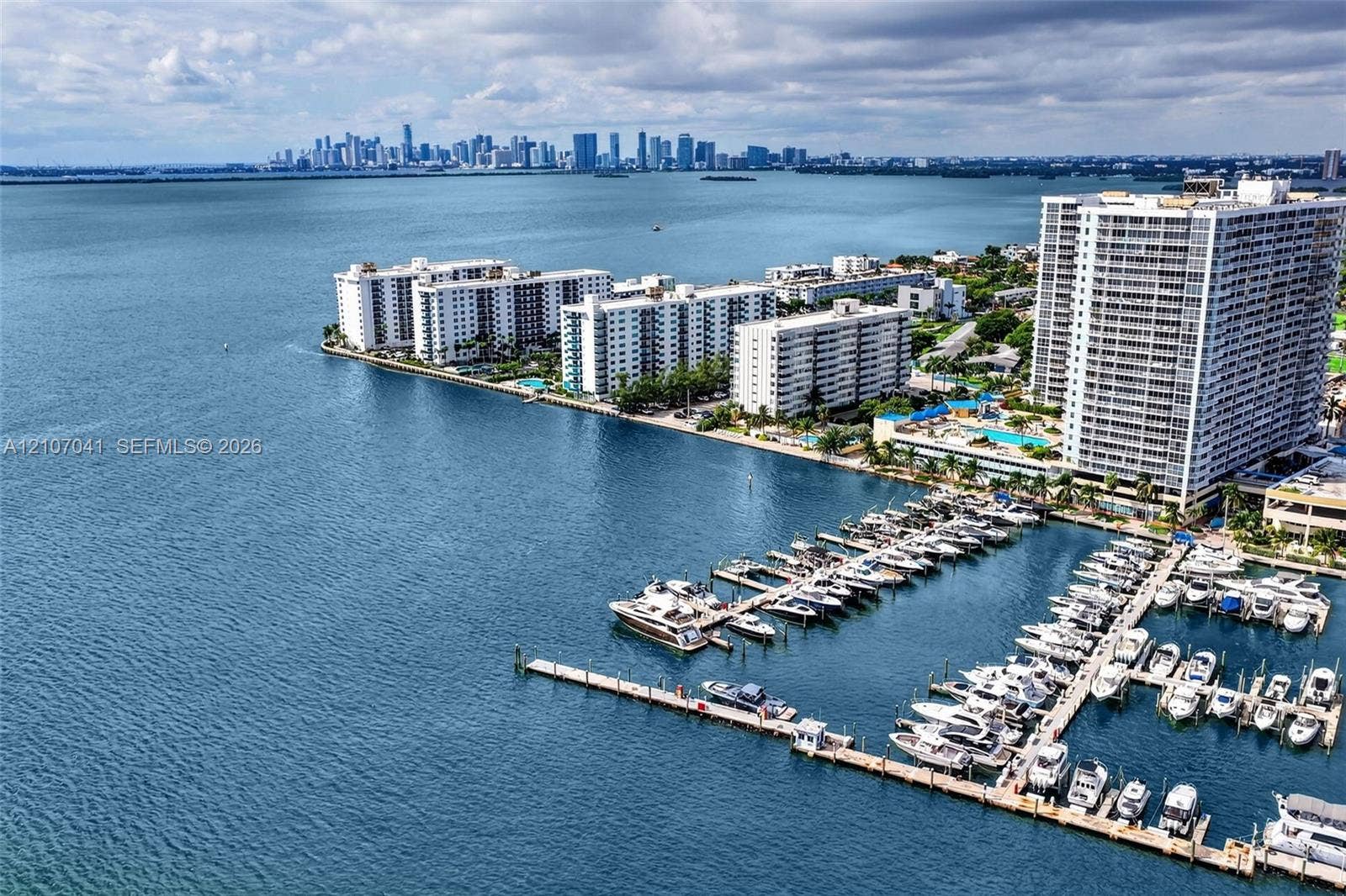 Aerial perspective of a waterfront residential complex featuring a private marina, deep-water boat slips, and a swimming pool, with distant urban skyline scenery