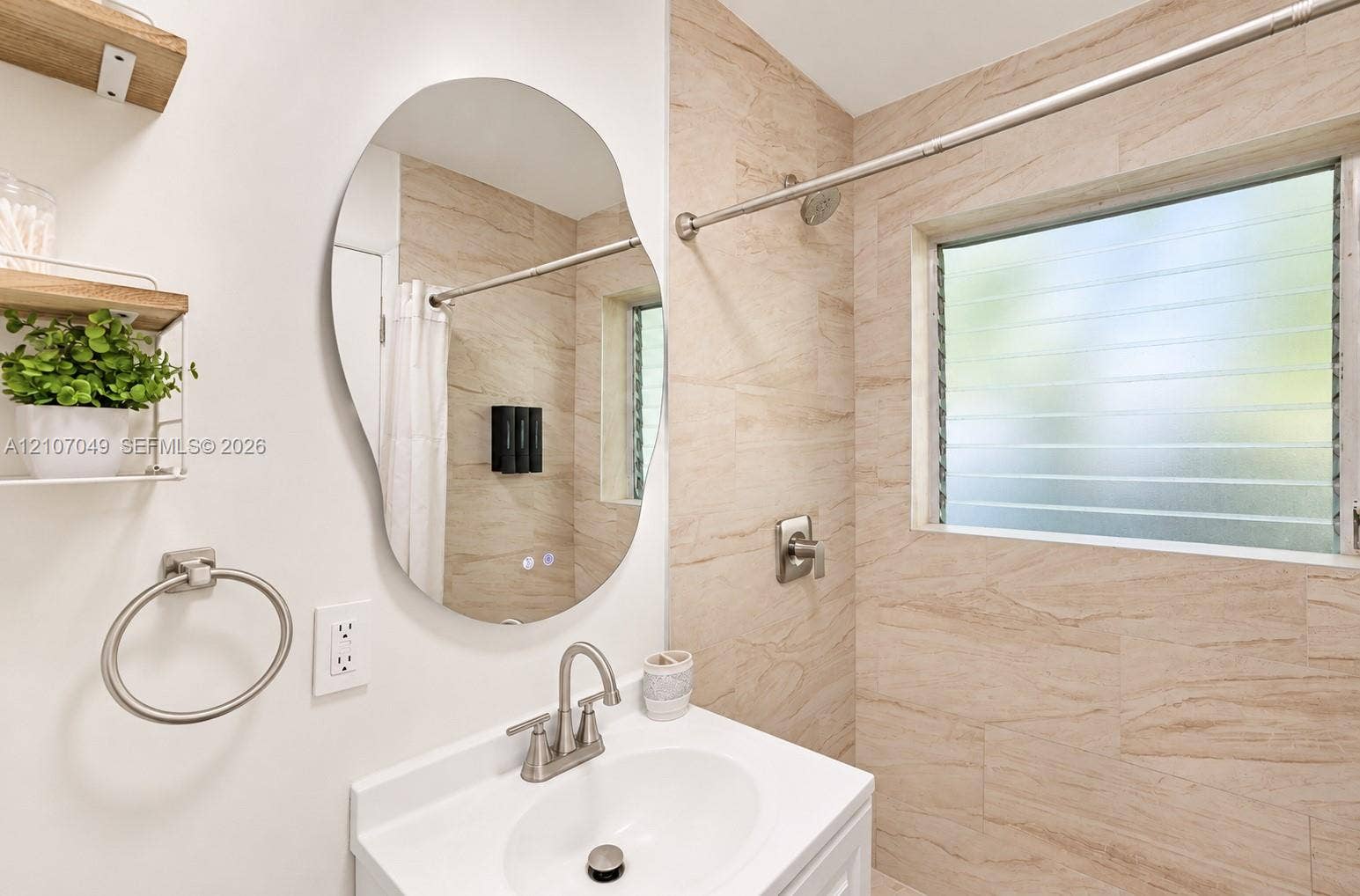 1009 NE 18th Street, Fort Lauderdale, FL 33305 Bathroom featuring a vanity with a white basin, an irregular-shaped wall mirror, louvered glass window, neutral-toned tiled shower surround, and satin nickel fixtures