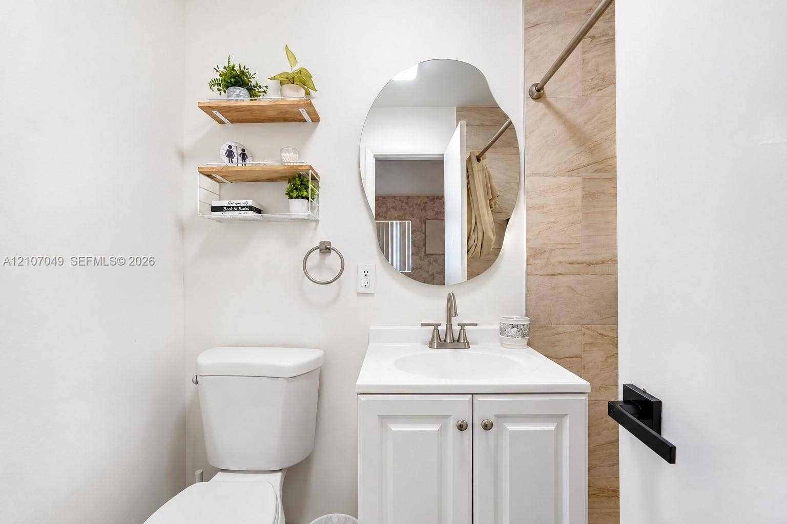 1009 NE 18th Street, Fort Lauderdale, FL 33305 White vanity with a solid surface countertop, organic-shaped wall mirror, two tiered wood-finish wall shelves, and neutral stone-finish tile surround
