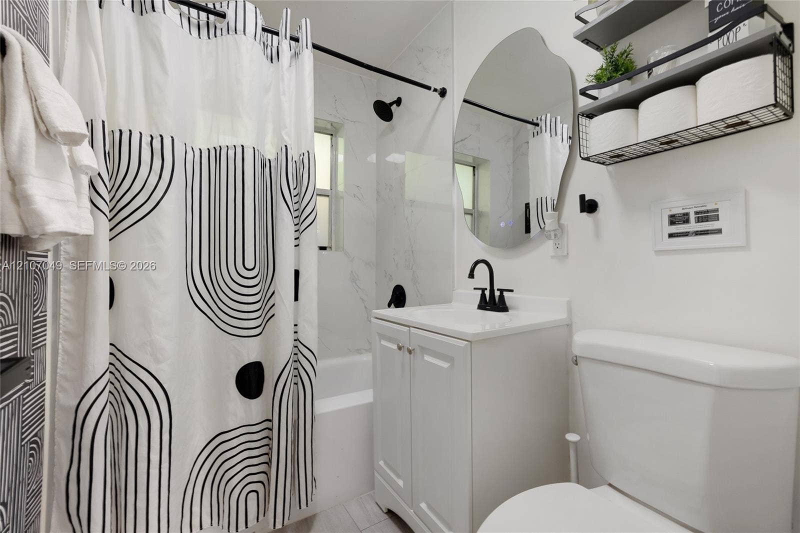 1009 NE 18th Street, Fort Lauderdale, FL 33305 Bathroom featuring a vanity with a white cabinet, stone-finish countertop, and matte black faucet, accompanied by a tub with light-toned wall tile and a matte black shower fixture, complemented by an irregular-shaped wall mirror