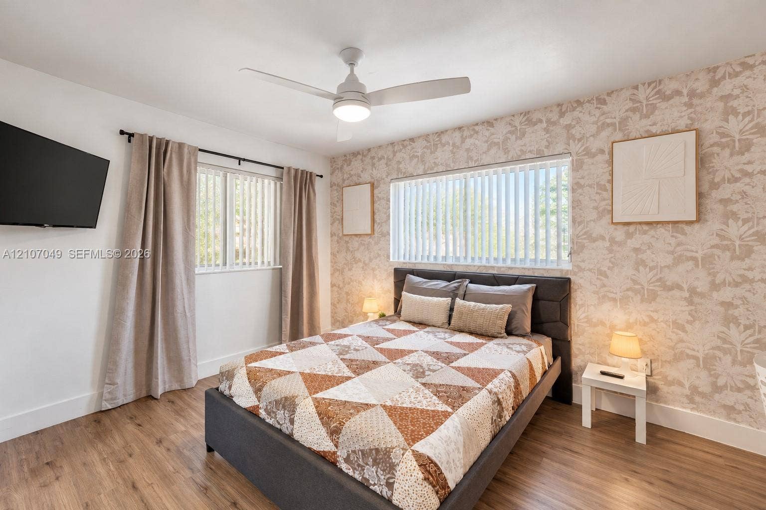 1009 NE 18th Street, Fort Lauderdale, FL 33305 Bedroom featuring wood-finish flooring, patterned wallpaper accent wall, dual windows with vertical blinds, and a central ceiling fan with integrated lighting