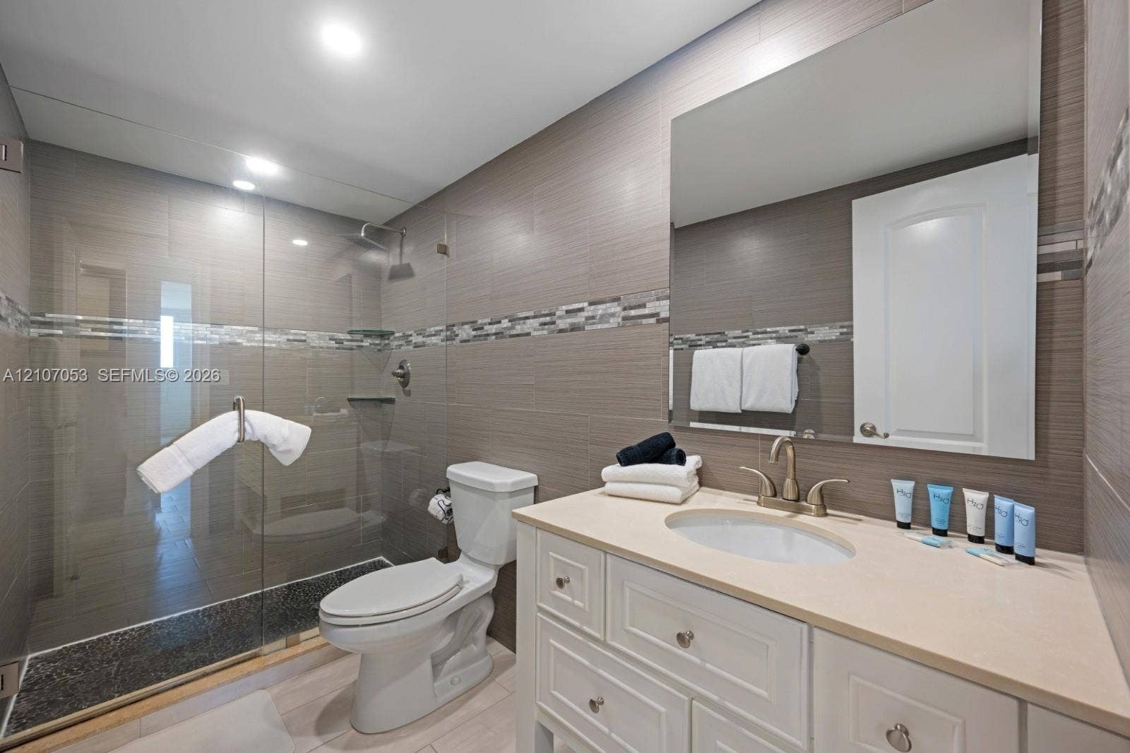 19370 Collins Avenue, #1403, Sunny Isles Beach, FL 33160 Bathroom featuring a white cabinetry vanity with a solid surface countertop, glass-enclosed shower with a decorative mosaic tile accent border, and full-width mirror