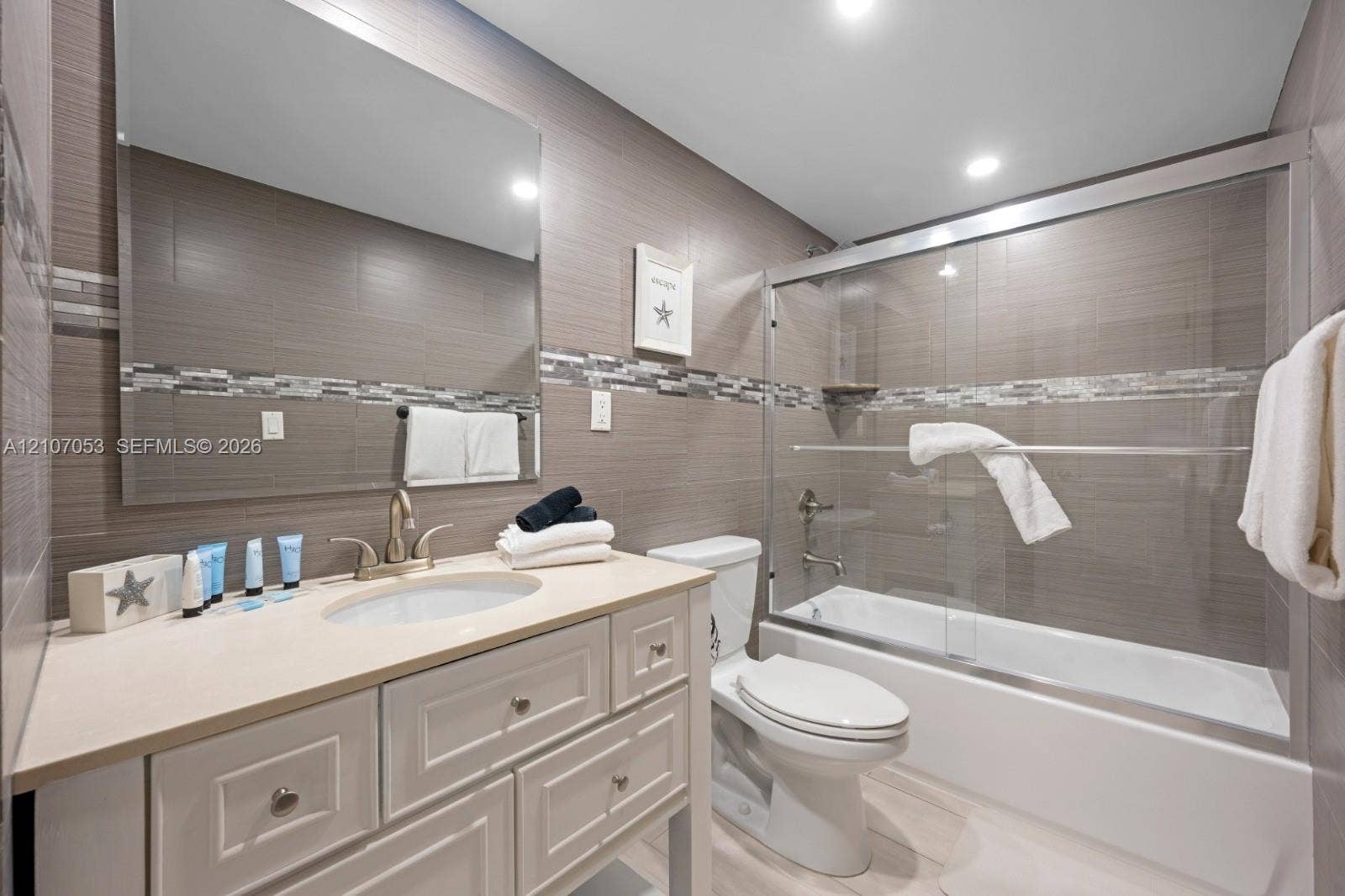 19370 Collins Avenue, #1403, Sunny Isles Beach, FL 33160 Bathroom featuring a white vanity cabinet with a light-toned countertop, an integrated oval basin, full-width mirror, tile-surround shower-tub combination with sliding glass doors, and horizontal decorative accent tiling