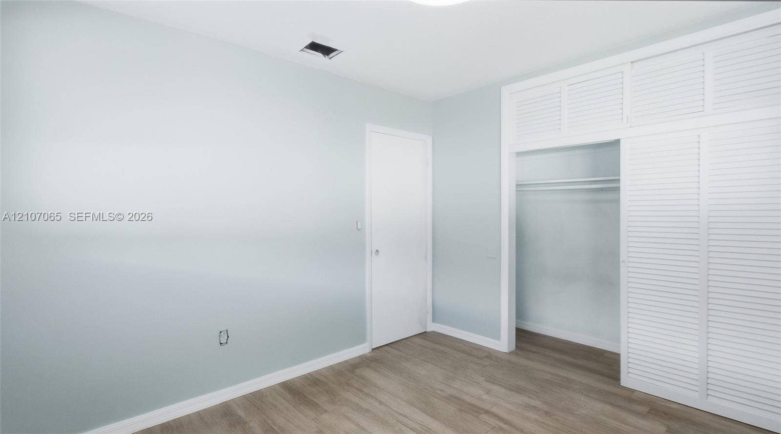 Room featuring wood-finish flooring, crisp neutral walls, and floor-to-ceiling built-in closet cabinetry with white louvered doors