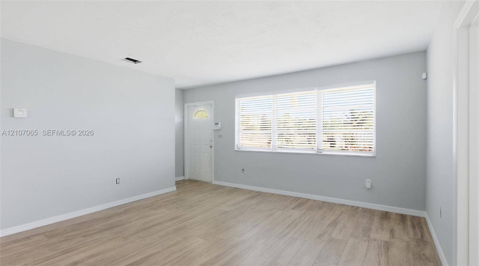 Neutral interior with light-toned wood-finish flooring, white walls, and a large triple-pane window with installed horizontal blinds