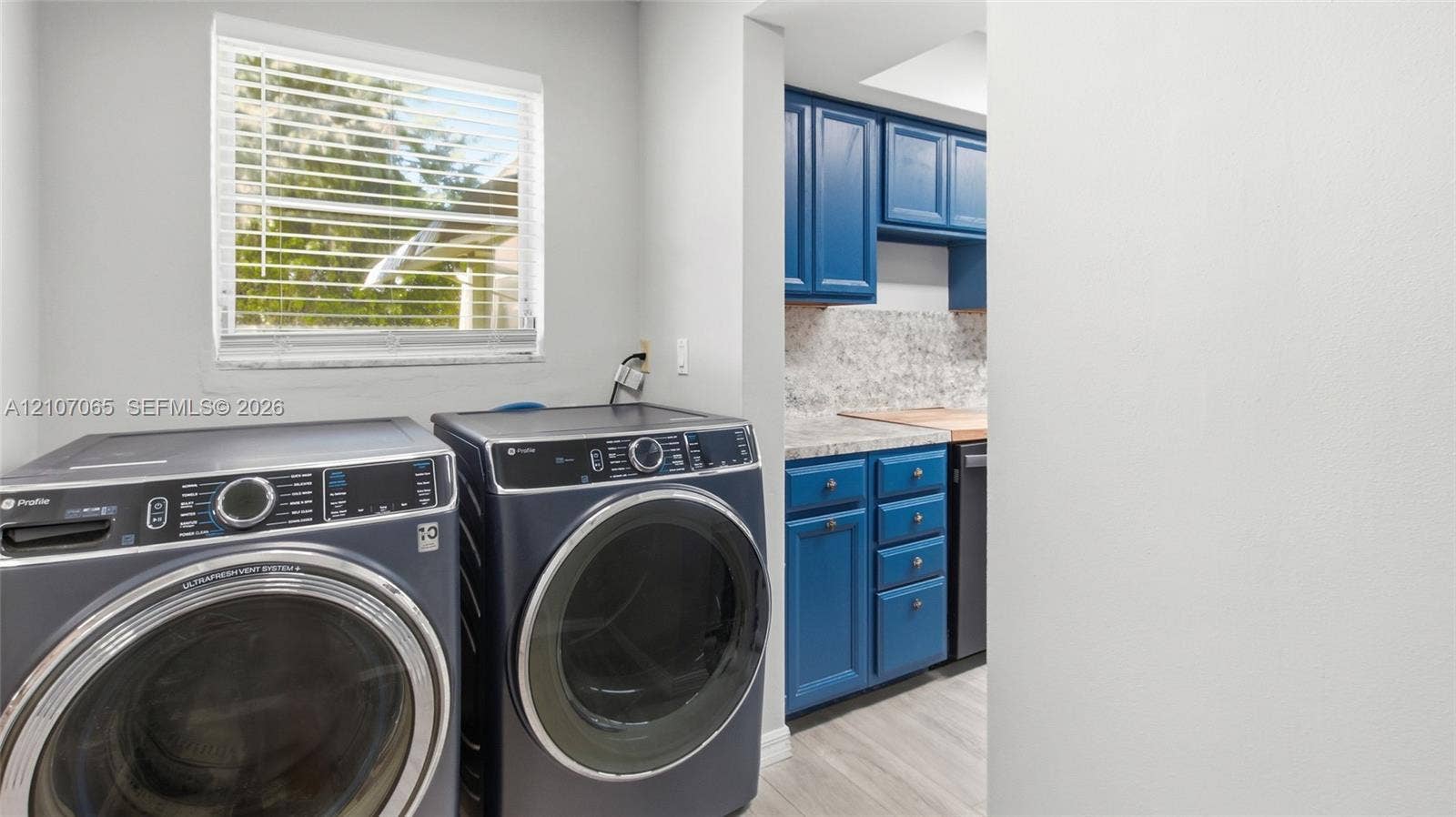 Laundry room featuring blue shaker-style cabinetry, stone-finish countertops, wood-finish flooring, and a window with horizontal blinds