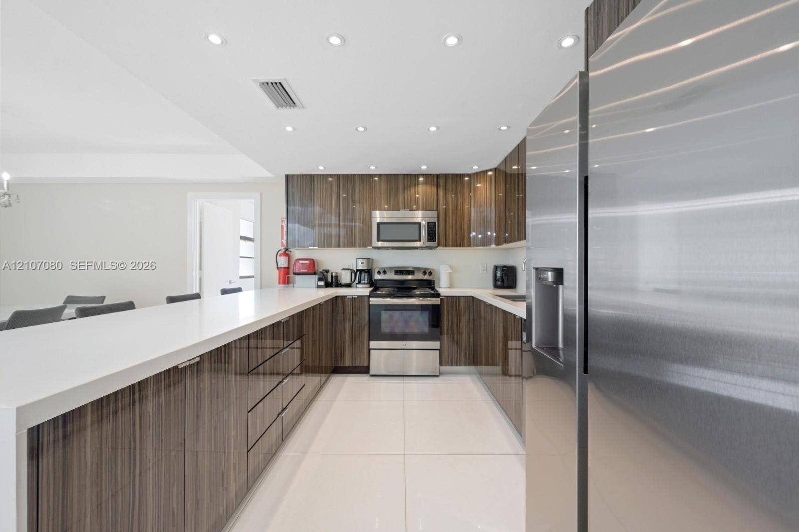 Kitchen featuring high-gloss wood-finish cabinetry, white solid-surface countertops, large-format tile flooring, stainless steel appliances, and recessed ceiling lighting