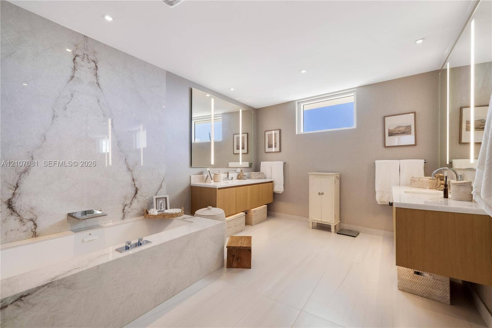 Bathroom featuring a soaking tub with stone-tiled surround, stone slab accent wall, dual floating wood-finish vanities, and vertical integrated mirror lighting