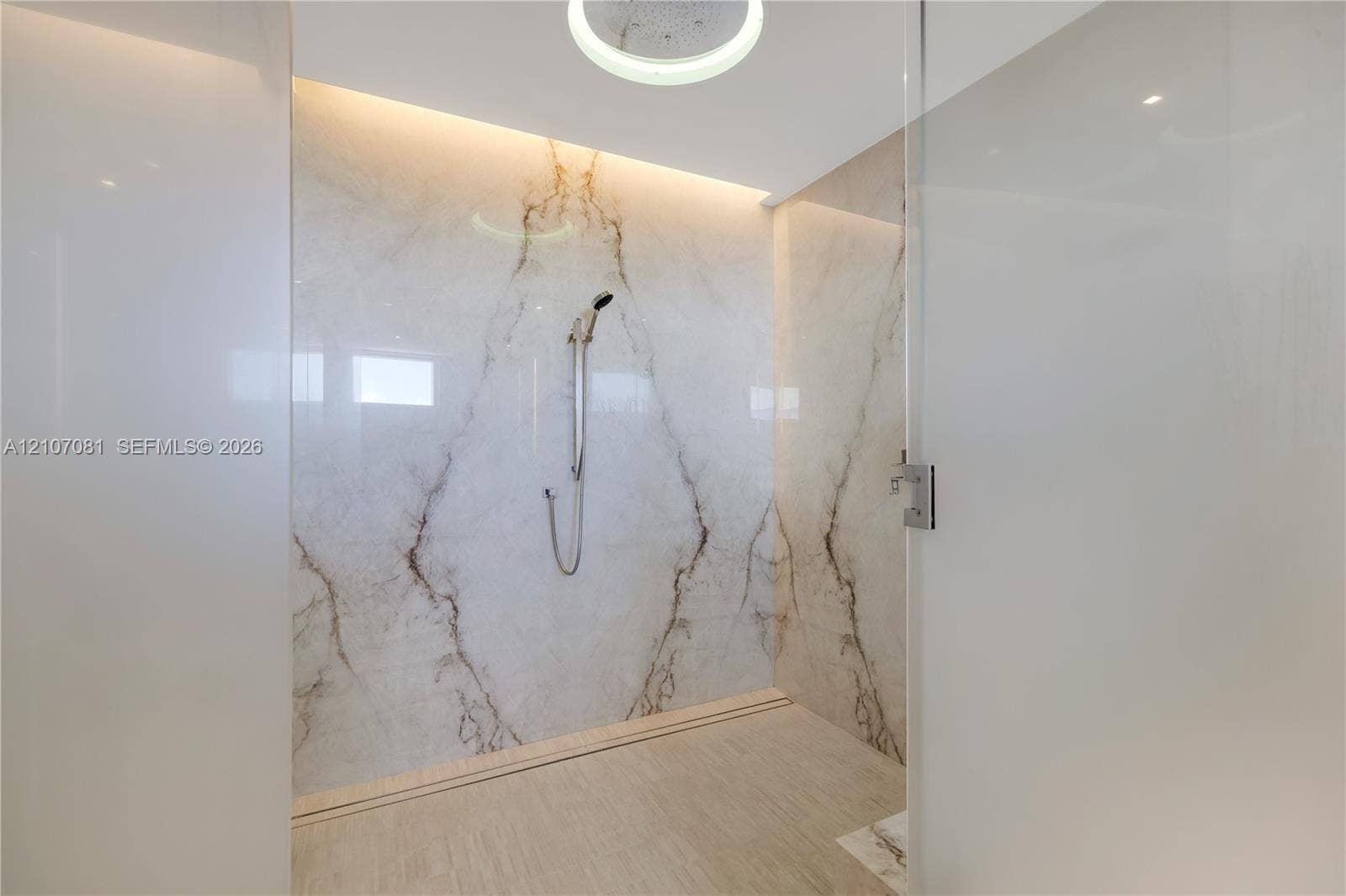 Walk-in shower enclosure featuring large-format veined stone wall panels, integrated ceiling lighting, a handheld shower fixture, and a glass door
