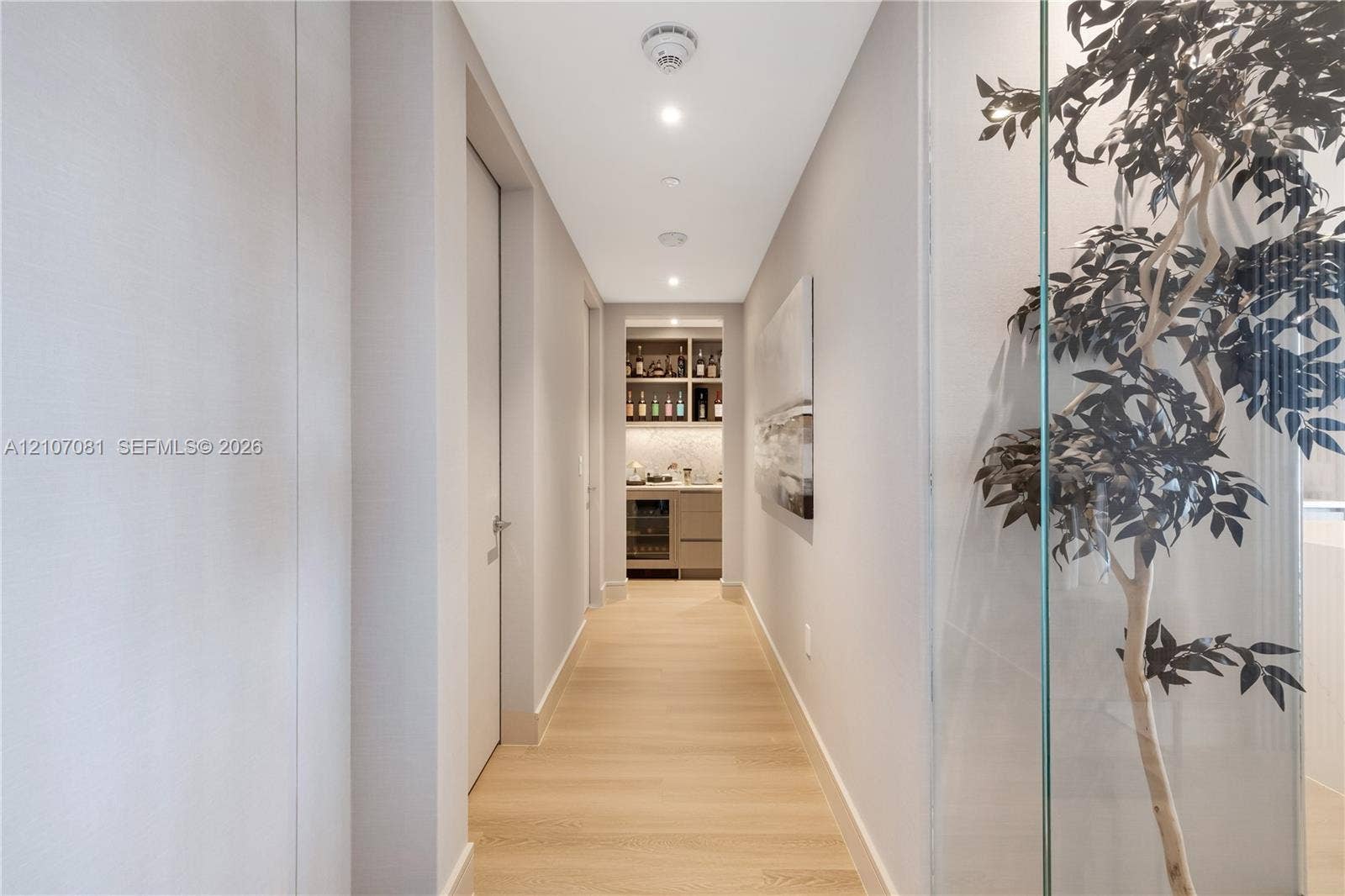 Hallway featuring light-toned wood-finish flooring, recessed lighting, and a glass-enclosed display area, leading to a built-in wet bar with cabinetry and stone-finish backsplash