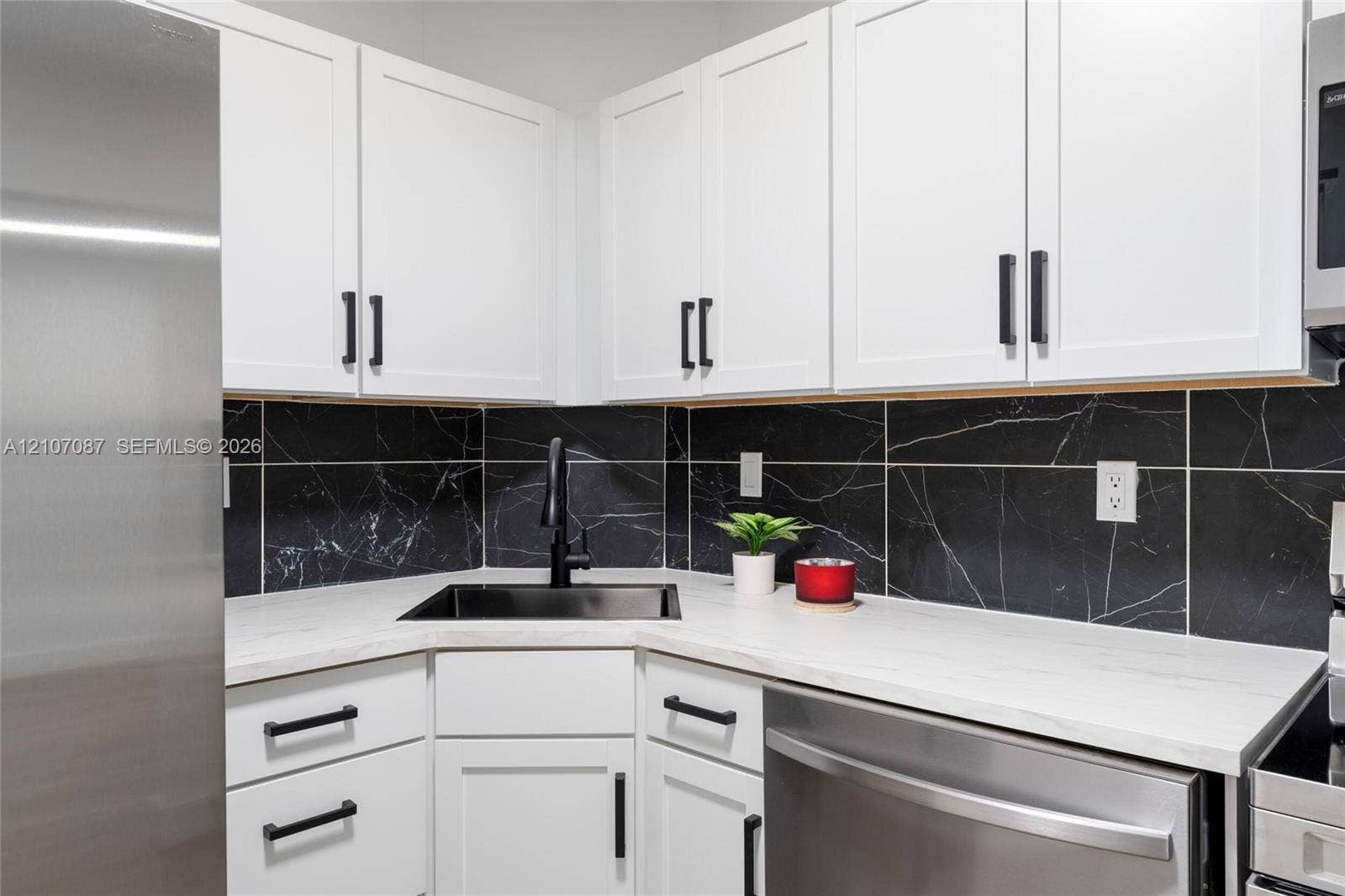 Kitchen featuring white shaker-style cabinetry, matte black hardware, black stone-patterned backsplash, white solid-surface countertops, and a stainless steel appliance suite