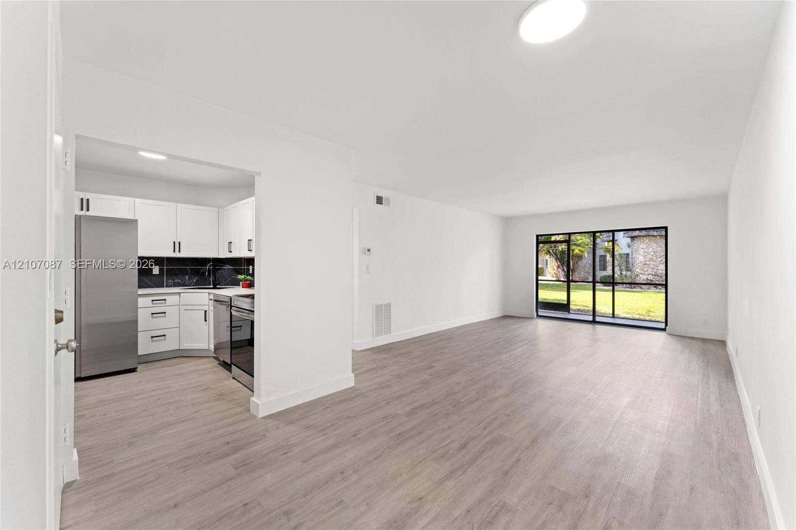 Kitchen featuring white shaker cabinetry, stainless steel appliances, and dark tile backsplash, complemented by wood-finish flooring and floor-to-ceiling glass sliding doors