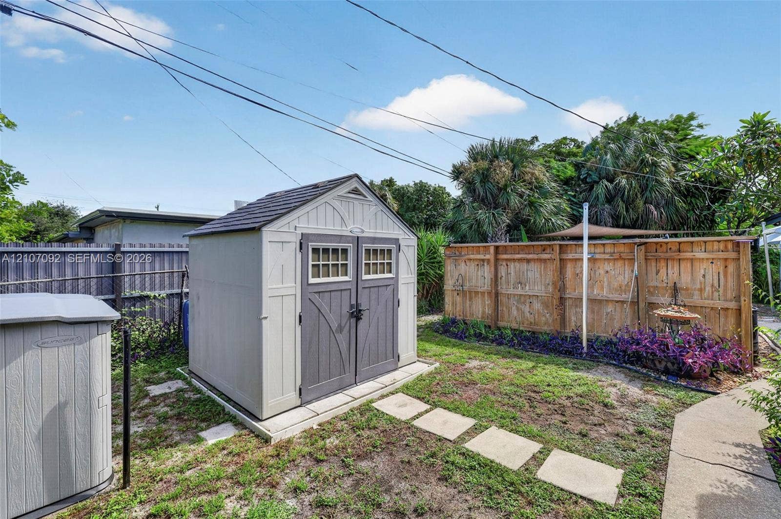 2864 NE 12th Terrace, Pompano Beach, FL 33064 Exterior storage shed featuring dual entry doors, shingle-style roof, and decorative window panes, complemented by a wood fence perimeter and stone paver walkway