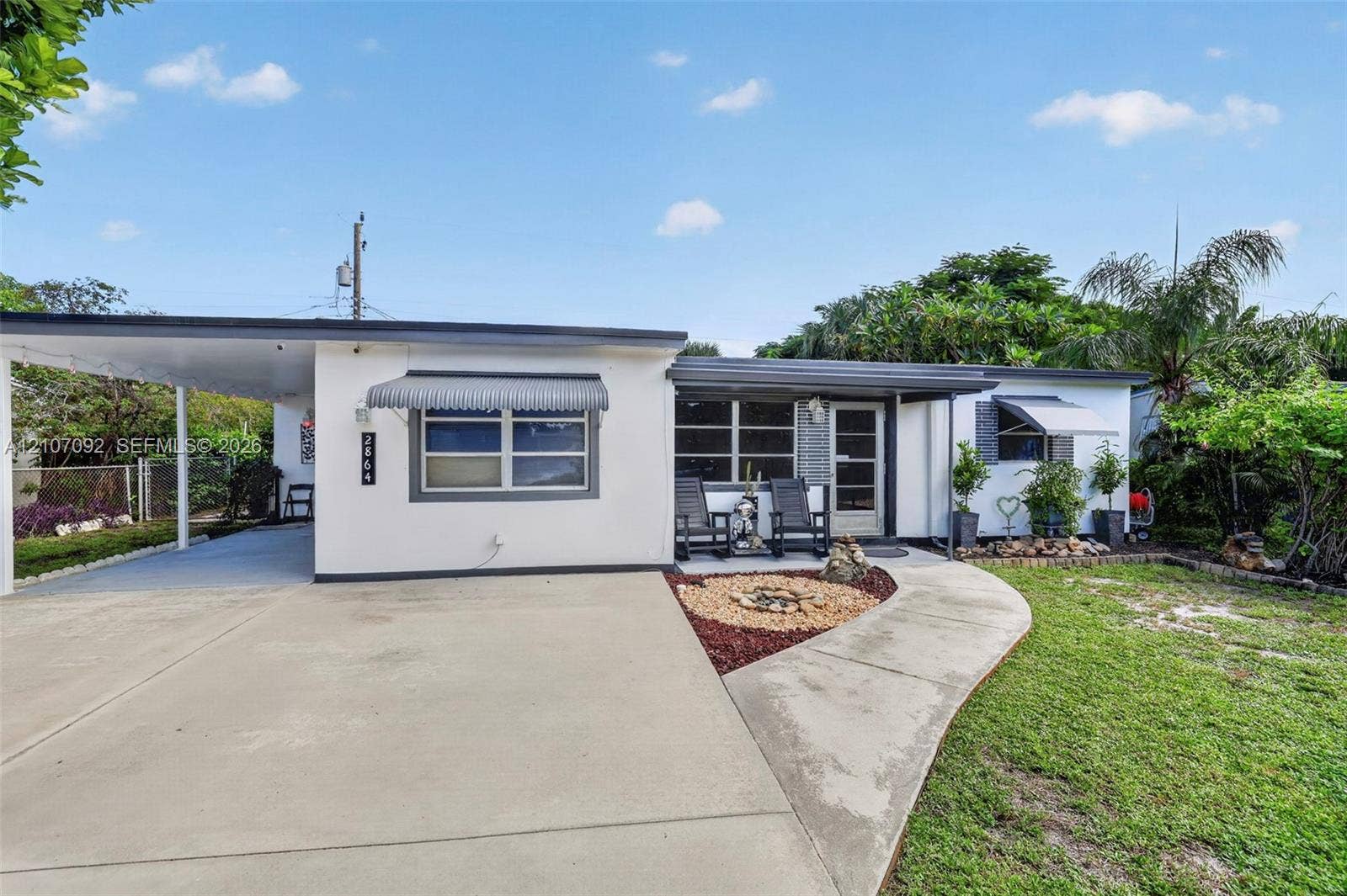 2864 NE 12th Terrace, Pompano Beach, FL 33064 Exterior featuring a covered carport, concrete driveway, metal window awnings, and a landscaped front yard with decorative stone mulch