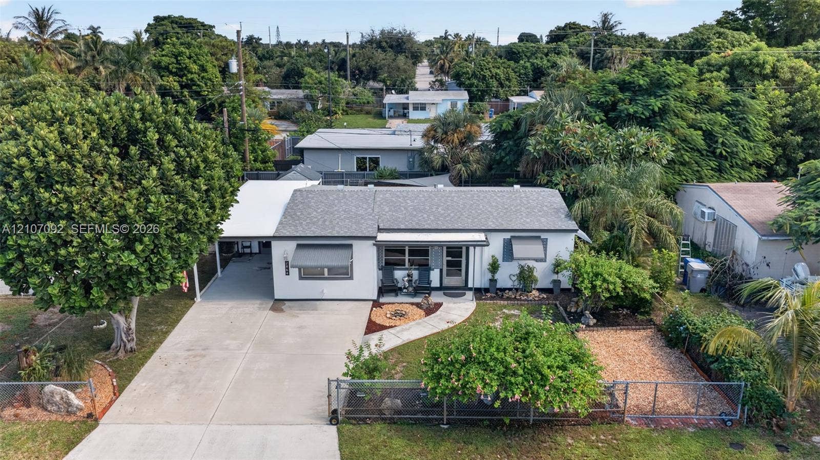 2864 NE 12th Terrace, Pompano Beach, FL 33064 Single-story exterior with light-toned siding, concrete driveway, attached covered carport, metal window awnings, and fenced front yard