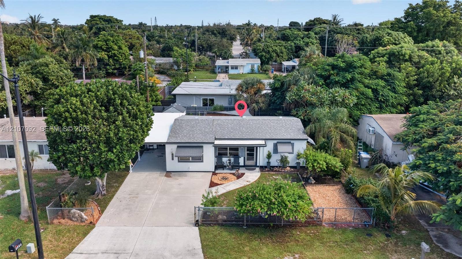 2864 NE 12th Terrace, Pompano Beach, FL 33064 Single-story residence featuring a concrete driveway, attached carport, metal awnings, fenced front yard, and mature landscaping