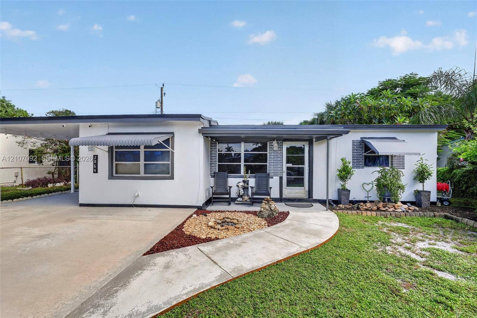2864 NE 12th Terrace, Pompano Beach, FL 33064 Mid-century modern exterior featuring a smooth stucco facade, metal-frame windows with retractable awnings, an attached carport, and a concrete walkway