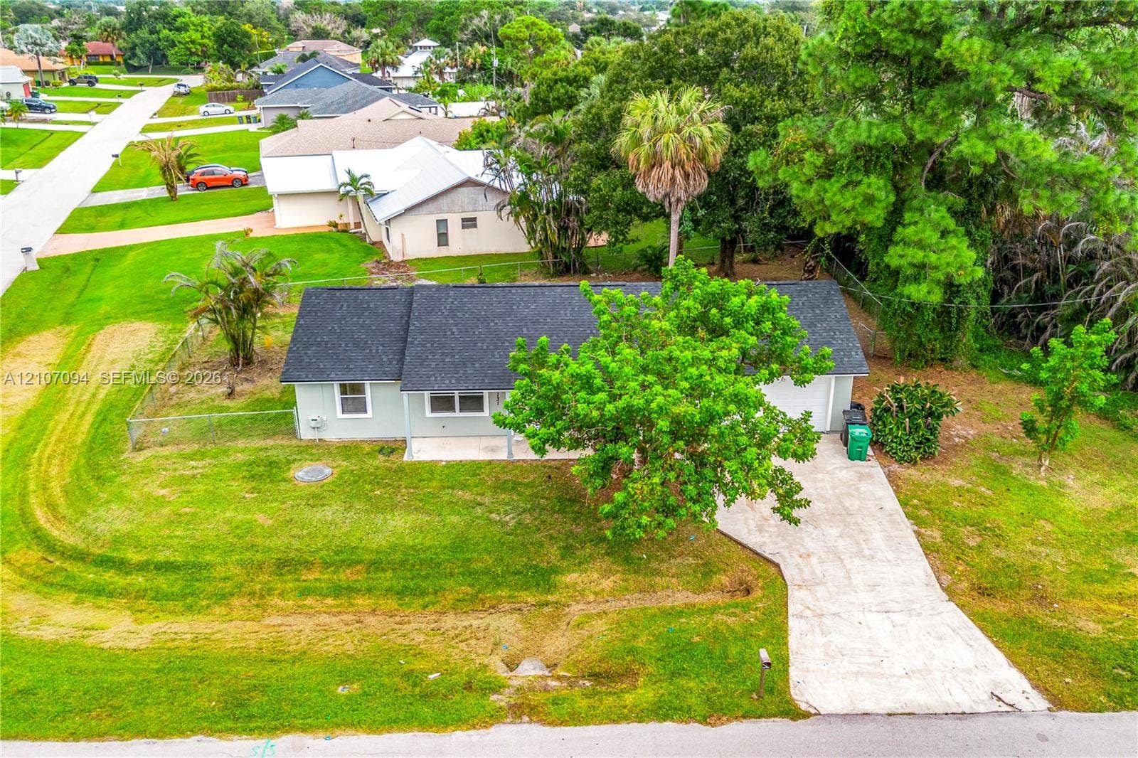 1131 SW London Lane, Port St Lucie, FL 34953 Single-story residence featuring a shingle roof, concrete driveway, and expansive lawn with mature trees