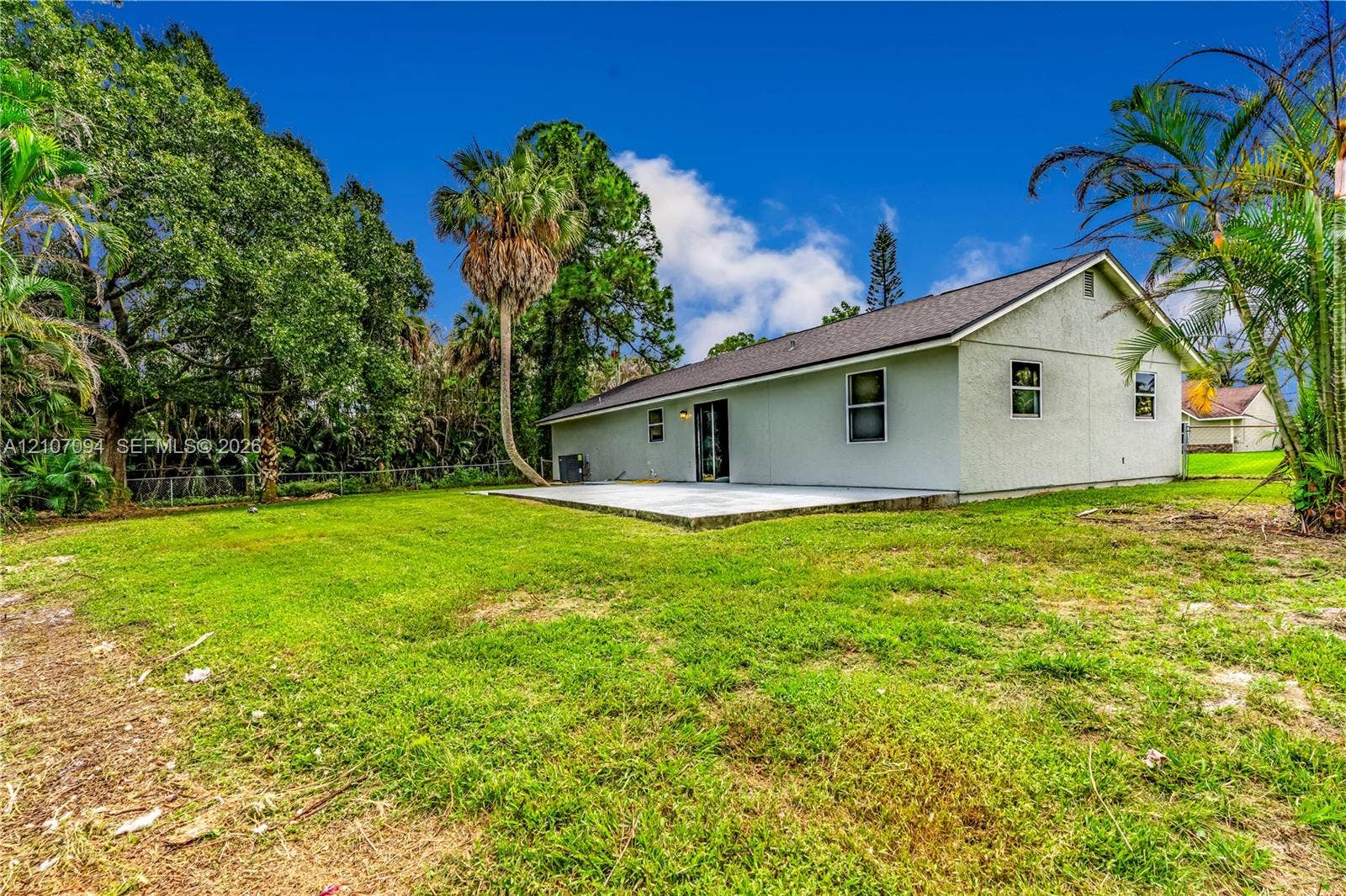 1131 SW London Lane, Port St Lucie, FL 34953 Exterior featuring a rear concrete patio, stucco siding, a pitched shingle roof, and a fenced backyard with mature trees