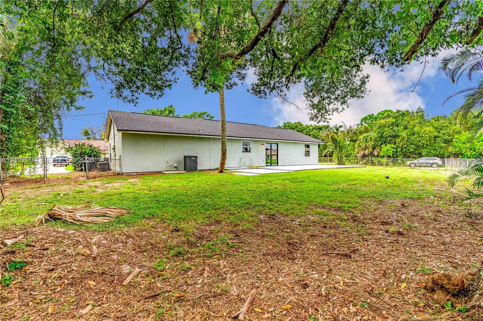 1131 SW London Lane, Port St Lucie, FL 34953 Backyard featuring a concrete patio, light-colored stucco exterior, shingle roof, chain-link perimeter fencing, and mature trees