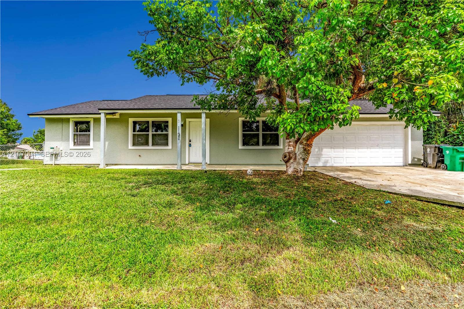 1131 SW London Lane, Port St Lucie, FL 34953 Single-story residence featuring a light-toned exterior, concrete driveway, attached garage, covered front porch, and expansive lawn
