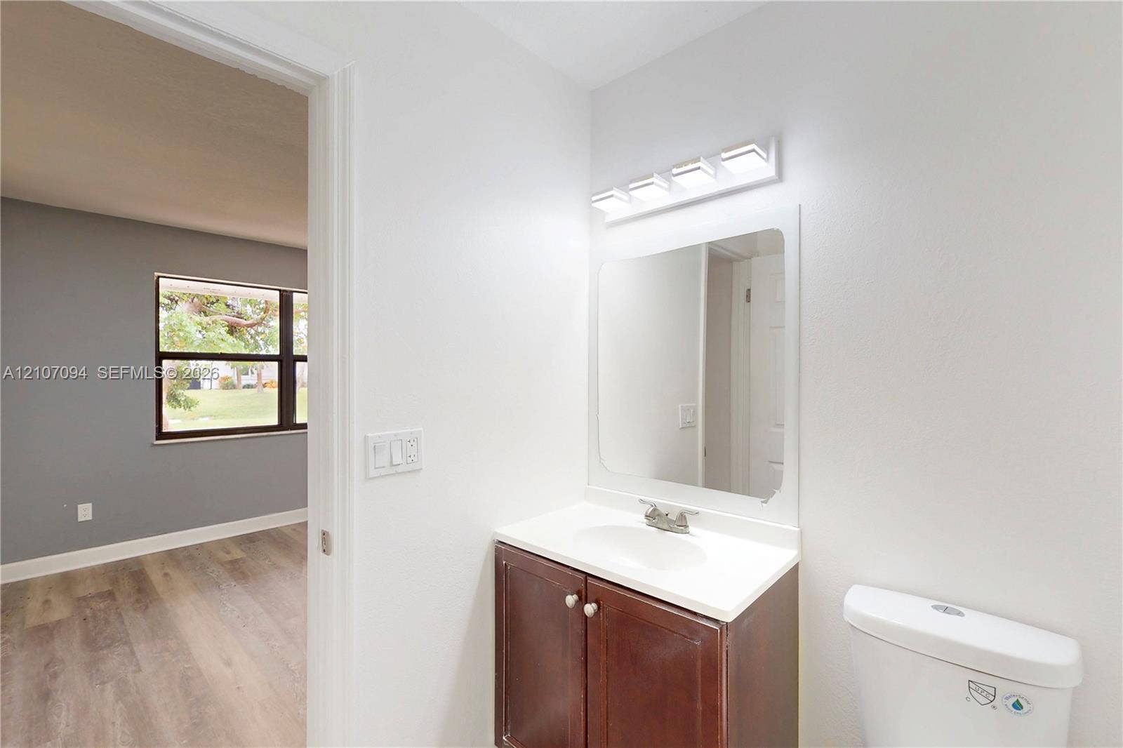 1131 SW London Lane, Port St Lucie, FL 34953 Vanity with integrated sink and countertop, polished chrome faucet, rectangular wall-mounted mirror, four-light vanity fixture, and wood-finish flooring