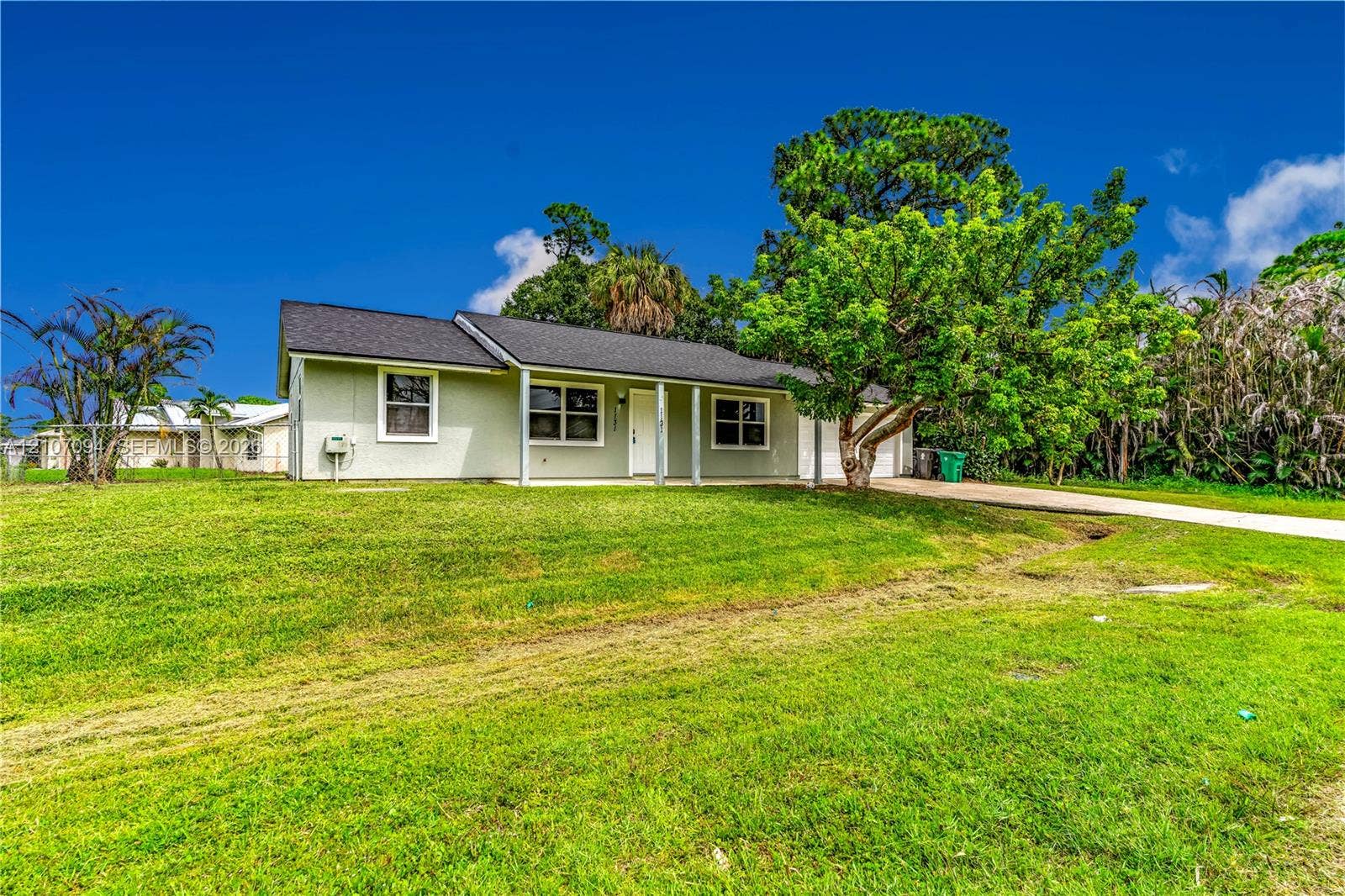 1131 SW London Lane, Port St Lucie, FL 34953 Single-story residence featuring a covered front porch, light-toned siding, dark shingled roof, and an expansive lawn