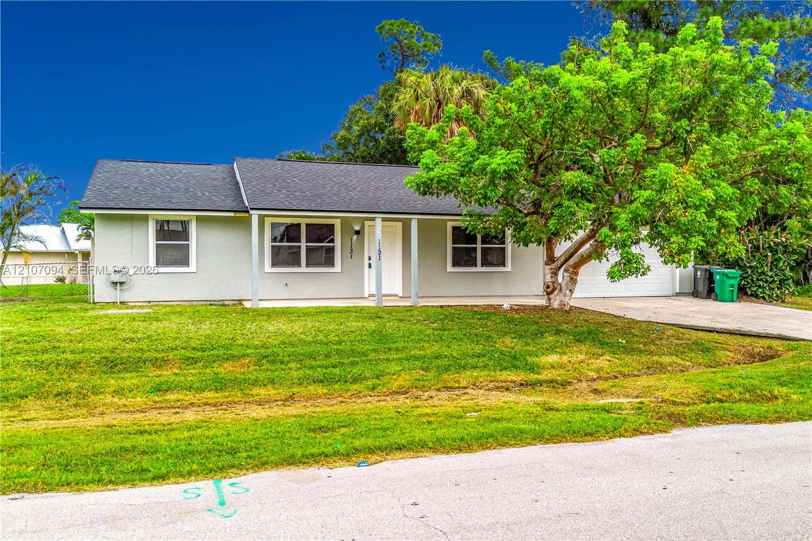 1131 SW London Lane, Port St Lucie, FL 34953 Single-story residence featuring a light-toned exterior, covered front porch, asphalt shingle roof, concrete driveway, and mature landscaping