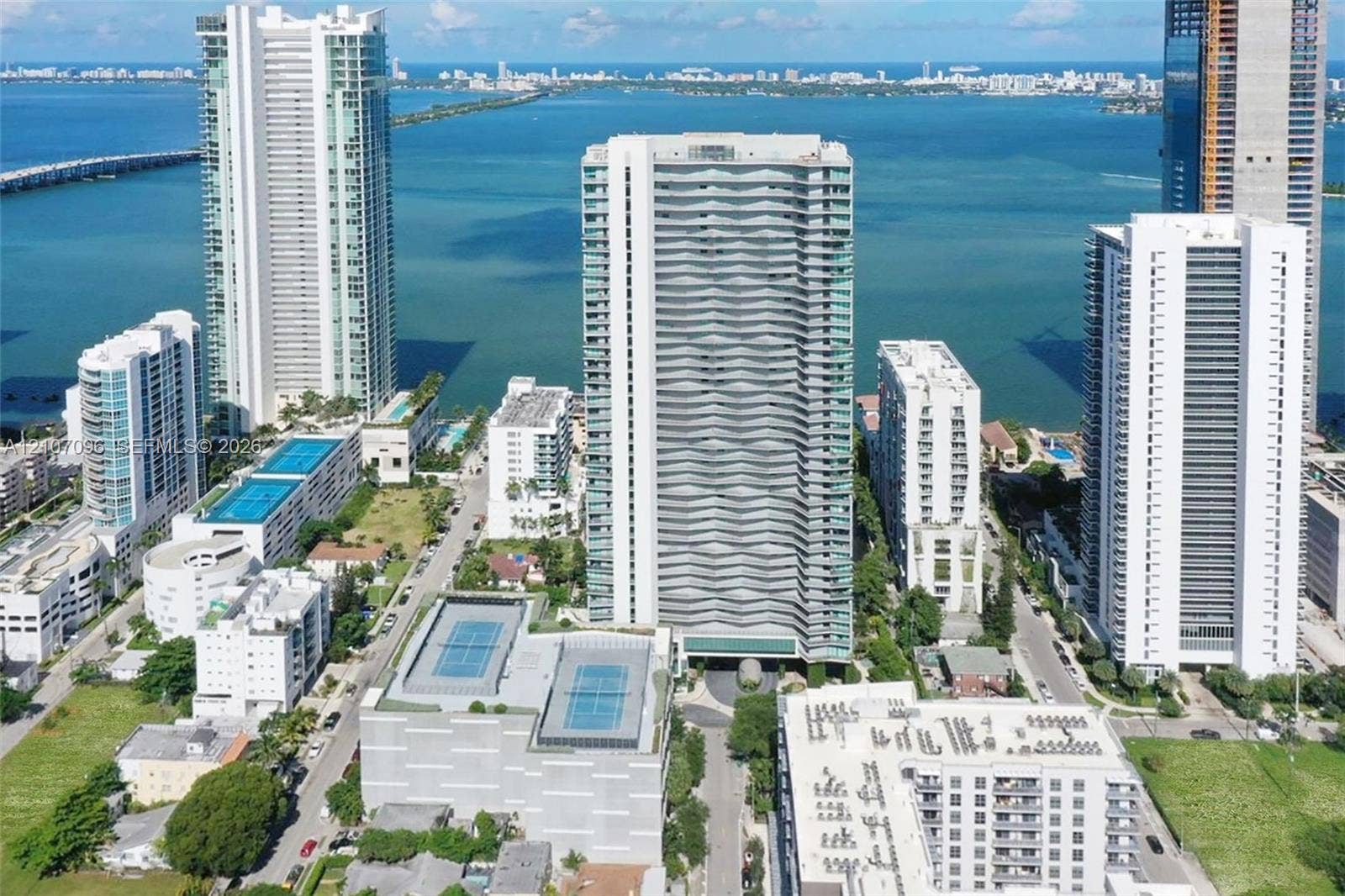 High-rise residential tower featuring a glass facade with balcony detailing, integrated parking structure with dual tennis courts, and direct coastal proximity