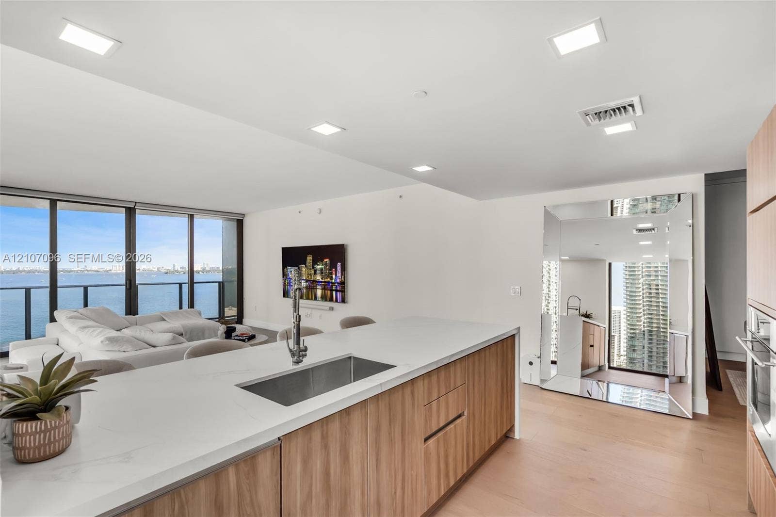 Kitchen island with solid surface countertop, undermount stainless steel sink, and wood-finish cabinetry, featuring floor-to-ceiling windows and recessed lighting
