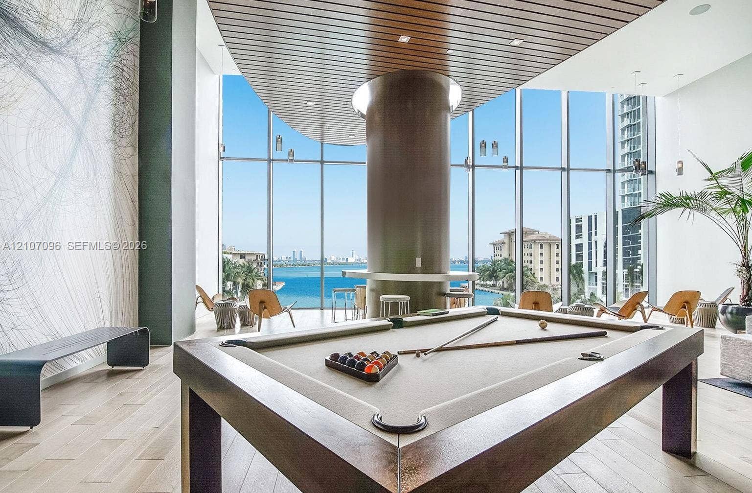 Floor-to-ceiling windows featuring panoramic water scenery, linear wood-slat ceiling detailing, hardwood flooring, and a central structural column with a built-in counter
