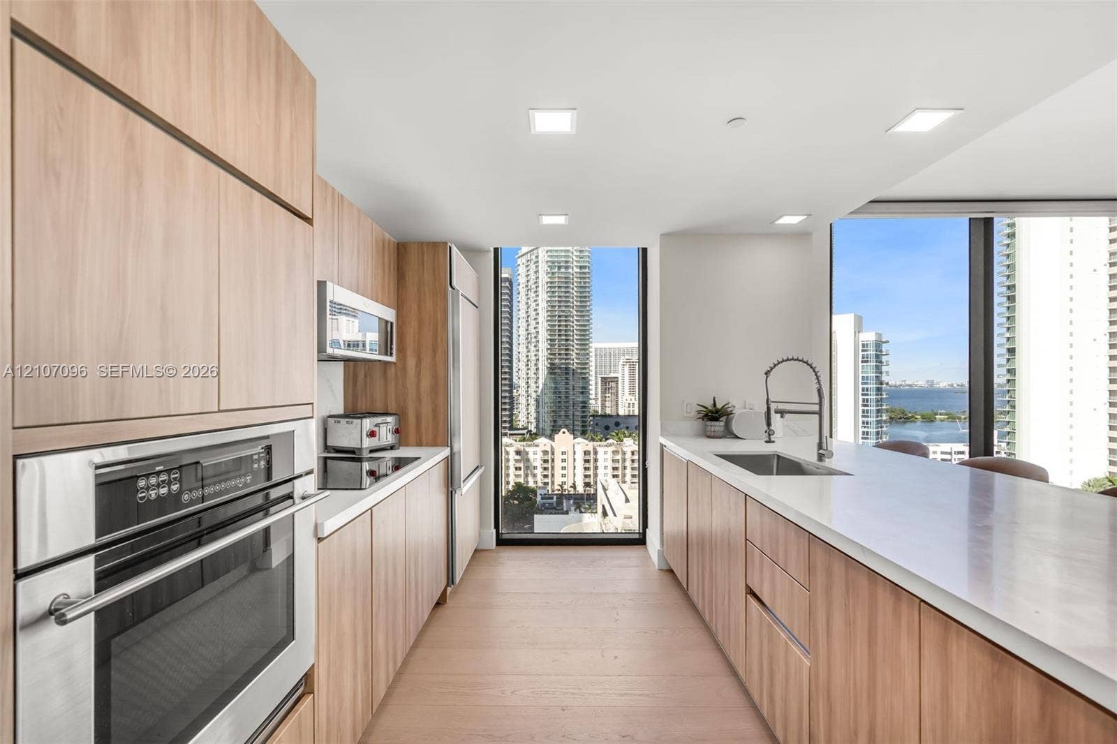 Kitchen featuring light wood-finish cabinetry, white solid-surface countertops, built-in stainless steel appliances, and floor-to-ceiling windows