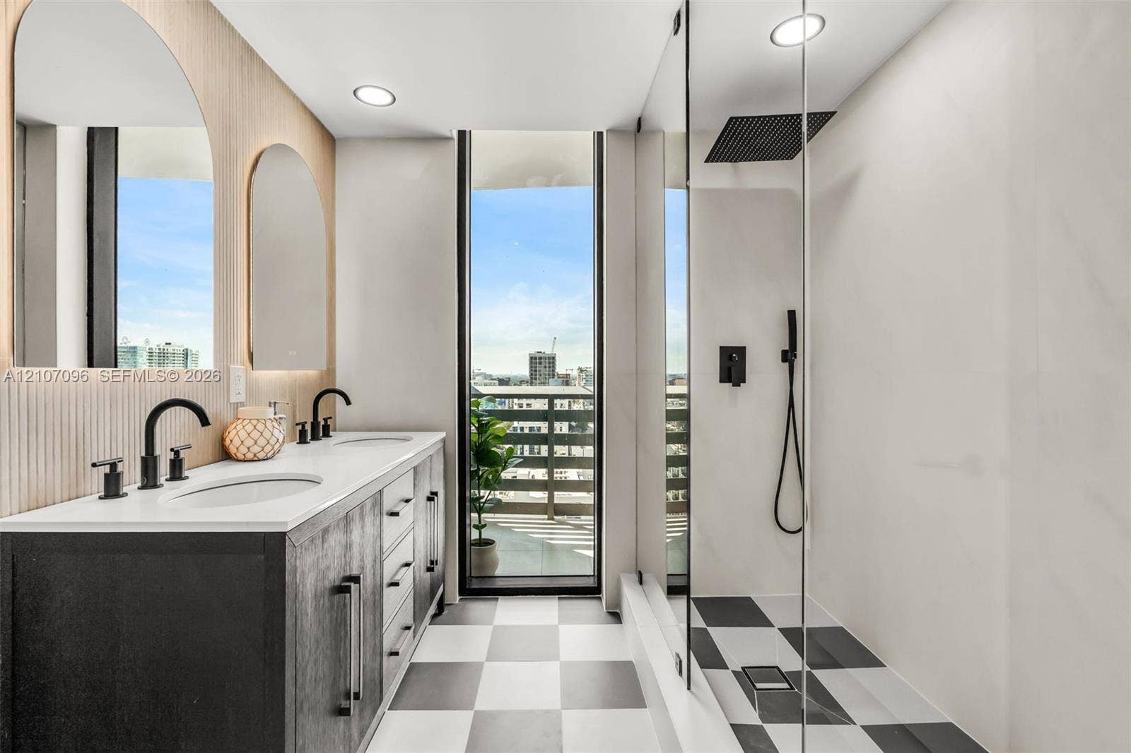 Double vanity with a white solid-surface countertop, dark cabinetry, arched wall mirrors, matte black fixtures, and a glass-enclosed shower featuring a rain-style showerhead and checkerboard tile flooring