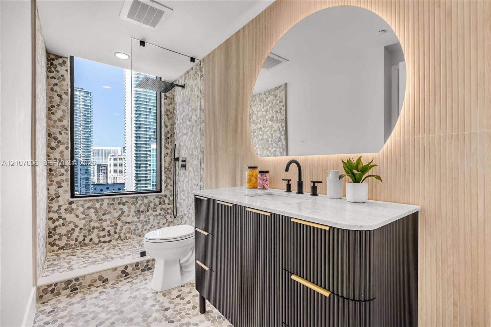 Custom dark fluted vanity with gold-tone hardware and light-toned stone countertop, integrated backlit arched mirror, floor-to-ceiling pebble tile shower surround, and matte black rain shower head