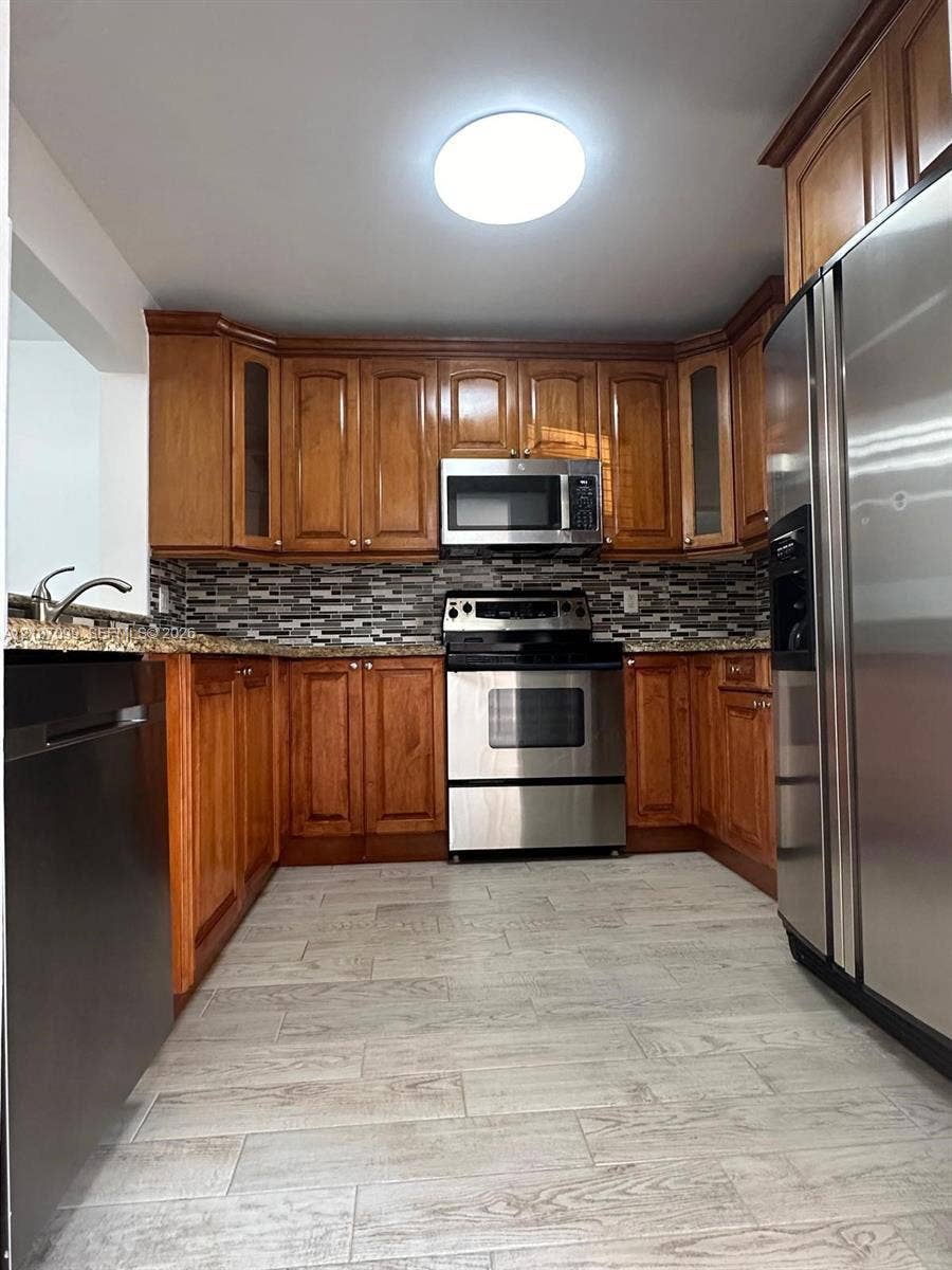 Kitchen featuring warm-toned wood cabinetry, multi-textured stone-look backsplash, stainless steel appliances, stone-finish countertops, and wood-finish flooring