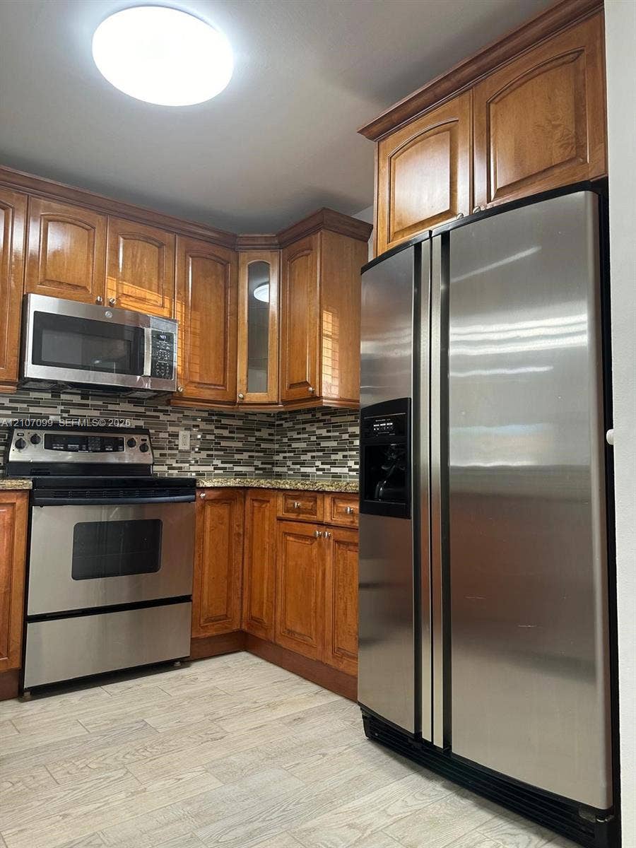 Kitchen featuring wood cabinetry, stainless steel appliances, stone-look countertops, a mosaic tile backsplash, and wood-finish flooring