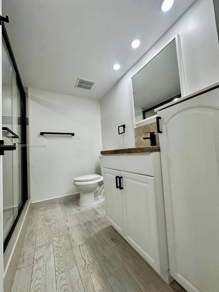 Bathroom featuring a vanity with stone-finish countertop, matte black hardware, recessed ceiling lighting, a walk-in shower with glass enclosure, and wood-finish flooring